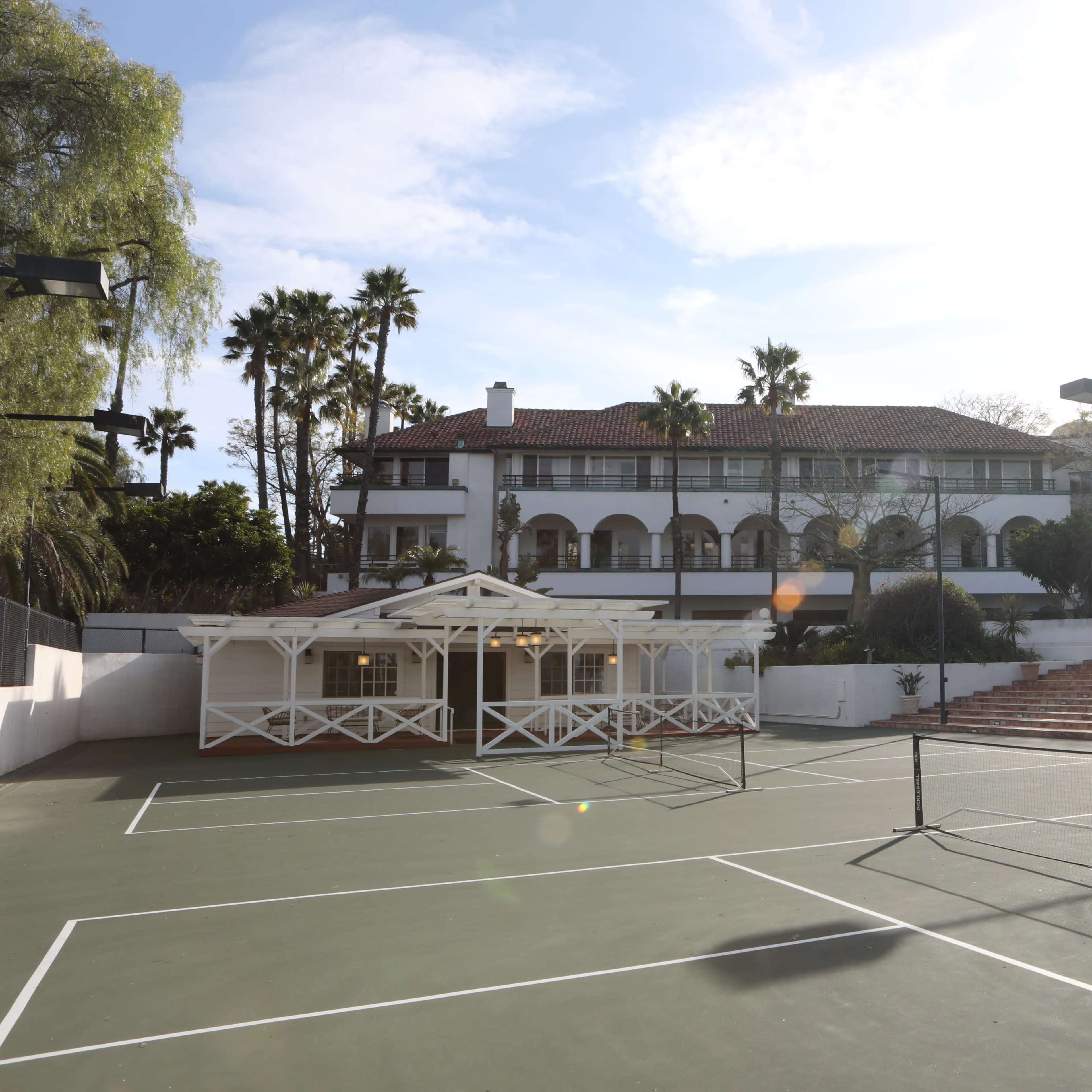 A tennis court with a view of a large building surrounded by palm trees under a bright sky.