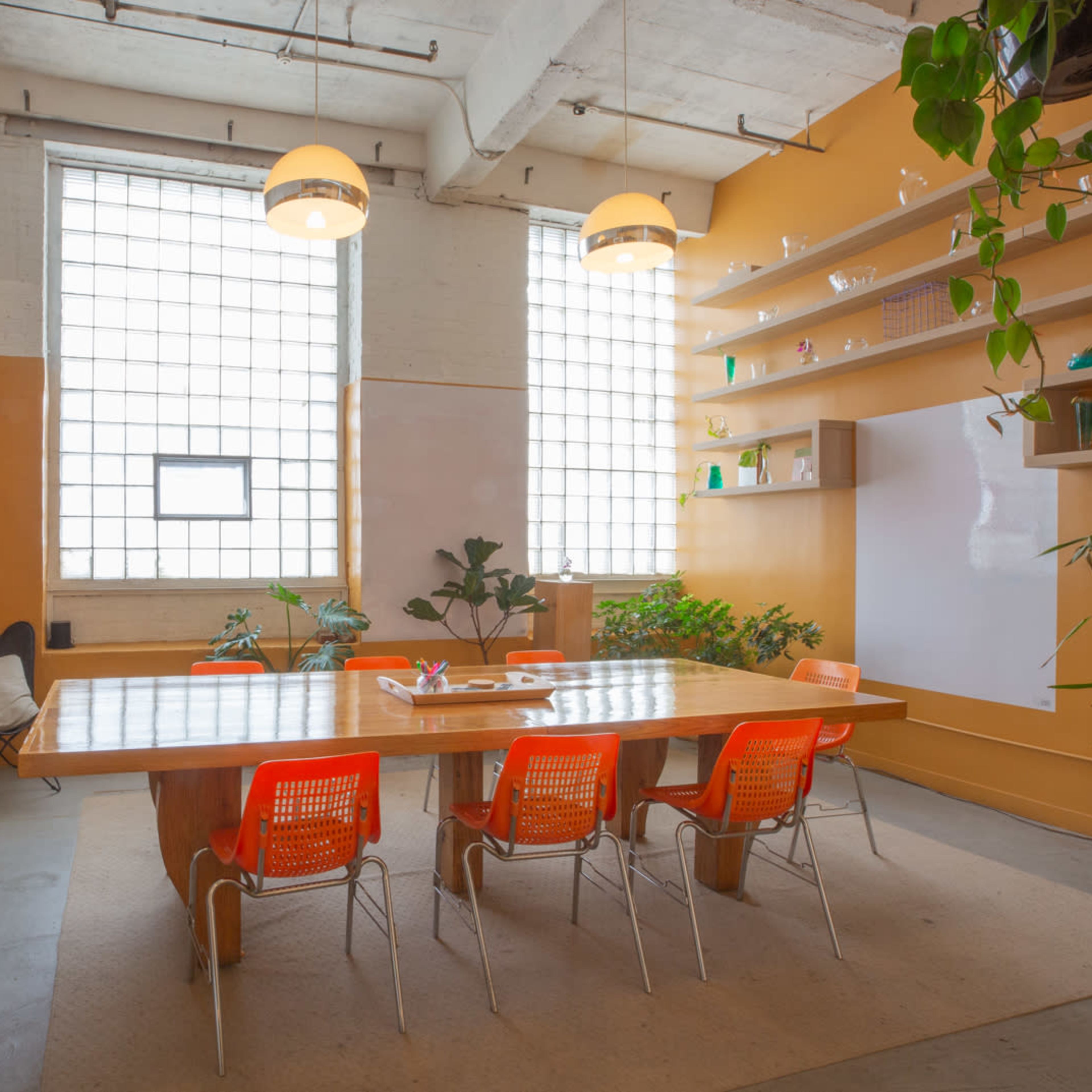 A bright meeting room features a large wooden table surrounded by six orange chairs, with plants and large windows providing natural light.