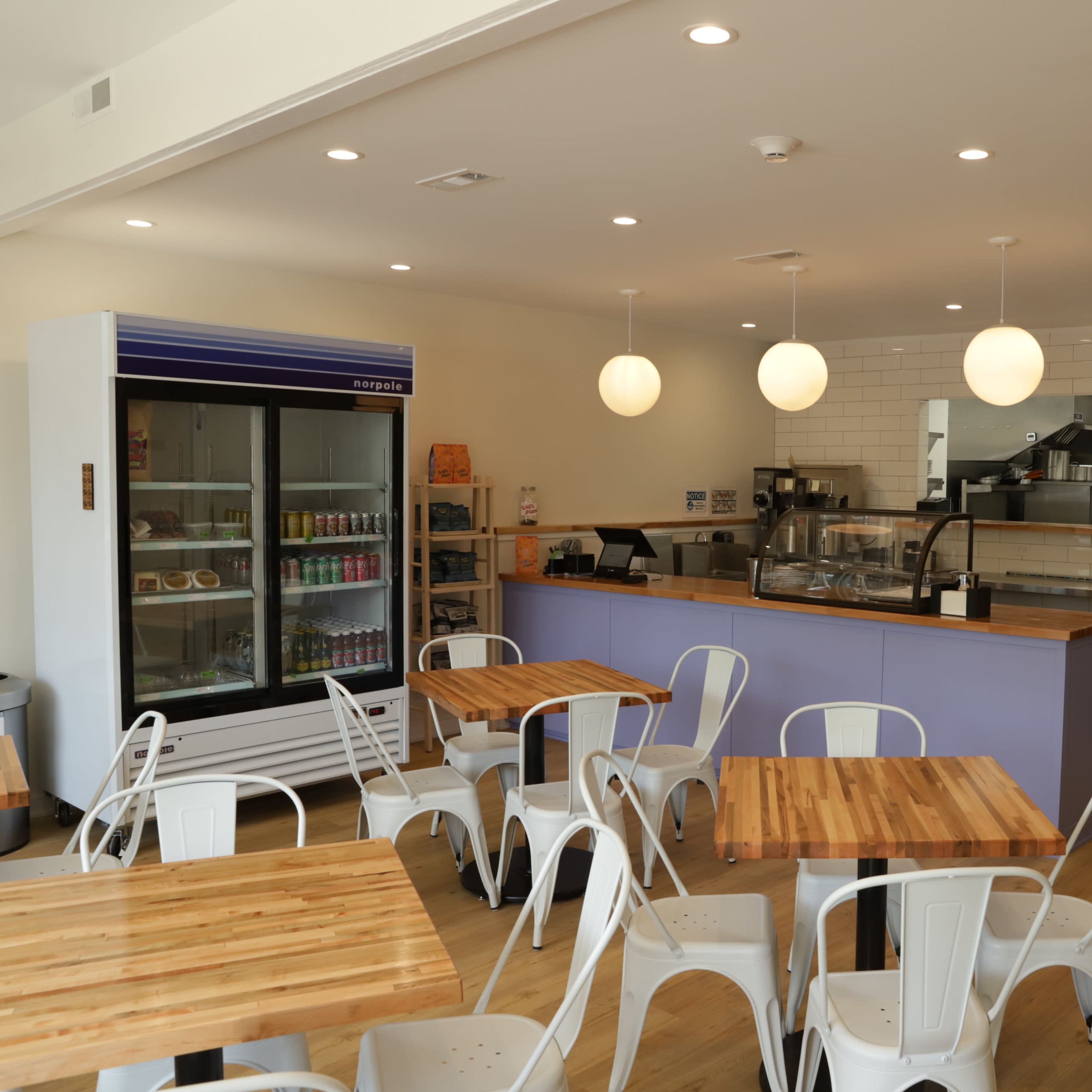 A modern café interior with wooden tables, white metal chairs, a service counter, and a refrigerator displaying beverages.