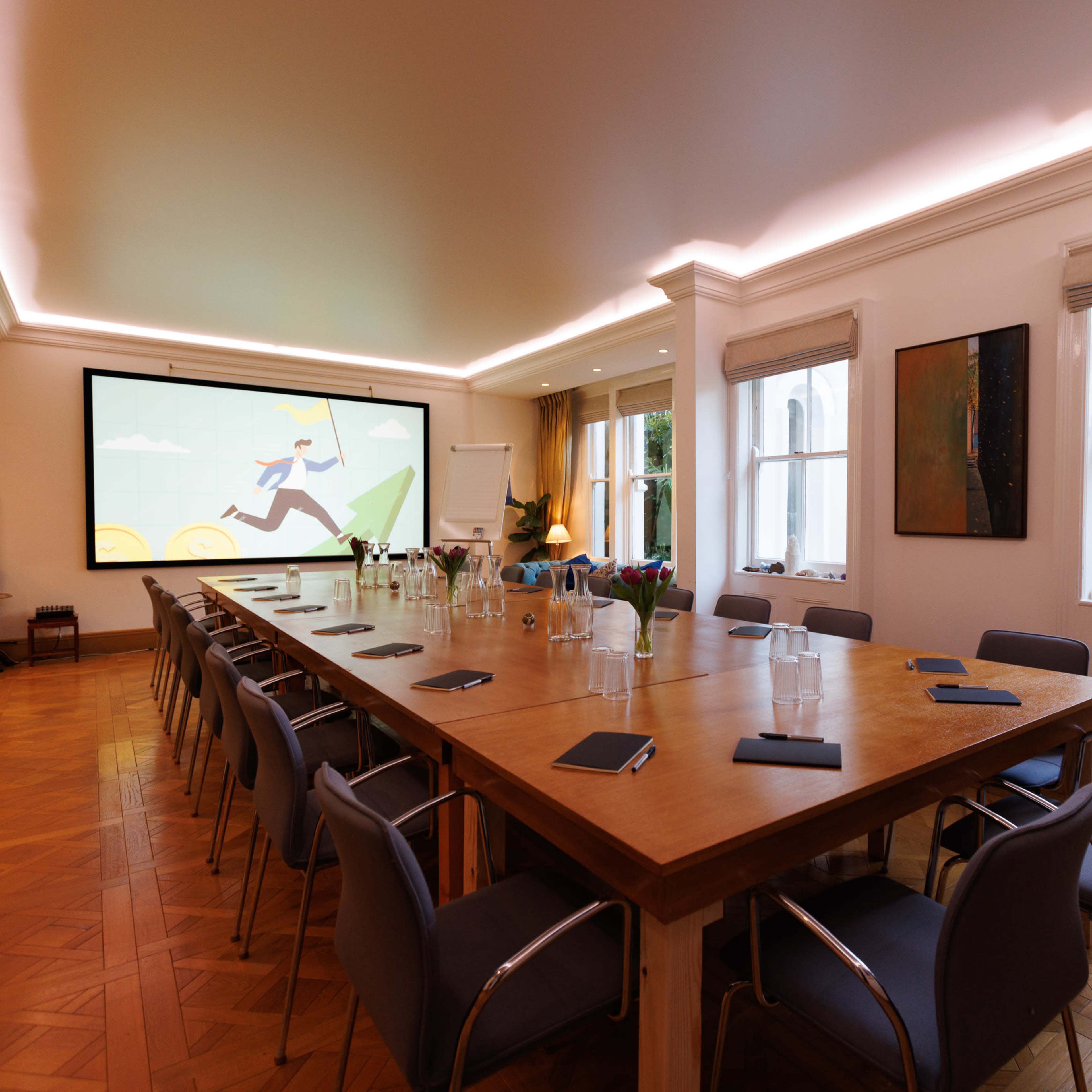 A large meeting room features a long wooden table, chairs, and a projector screen displaying a colorful graphic, with windows providing a view of an outdoor garden.