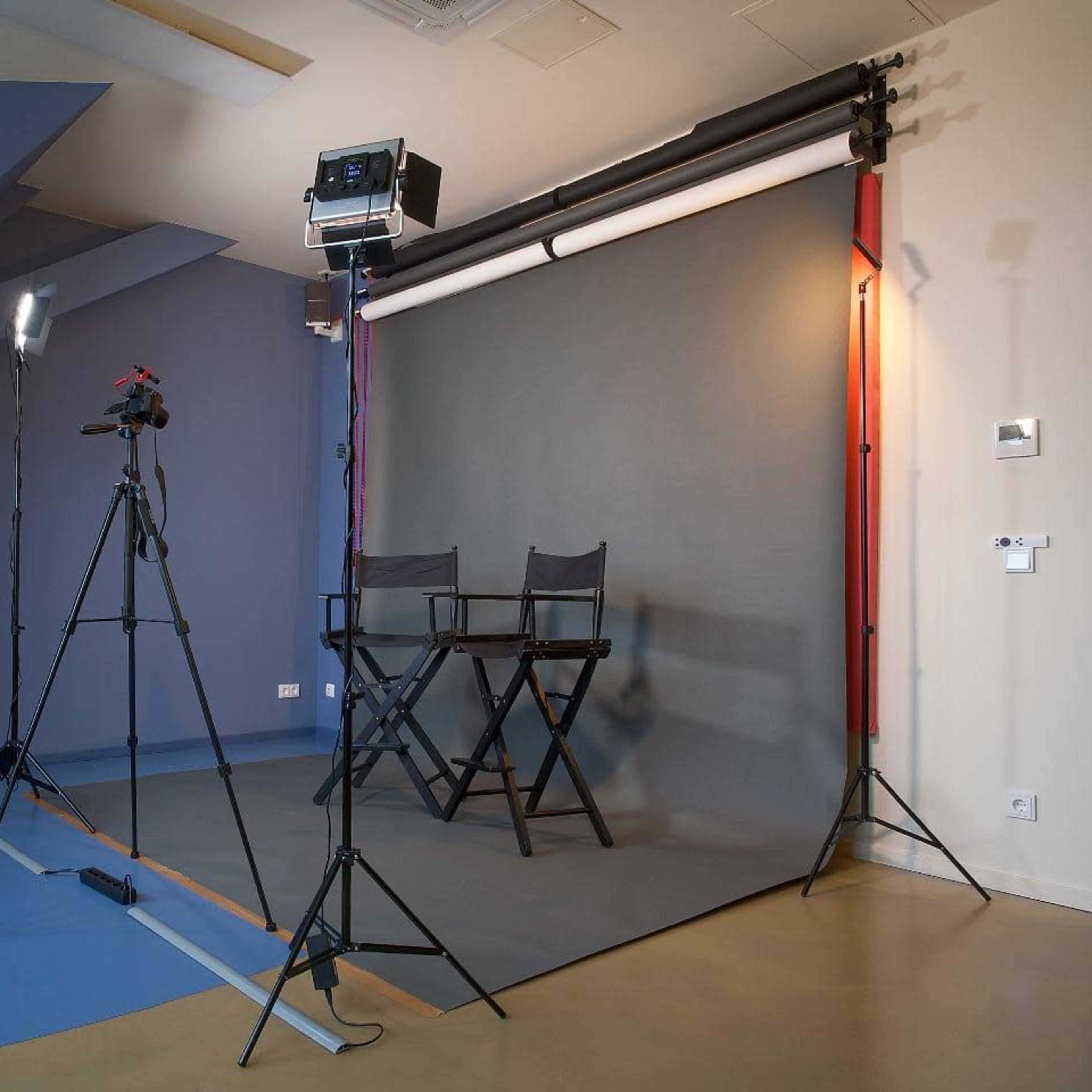 A video recording setup with a gray backdrop, two chairs, and multiple lighting and camera equipment in a studio environment.