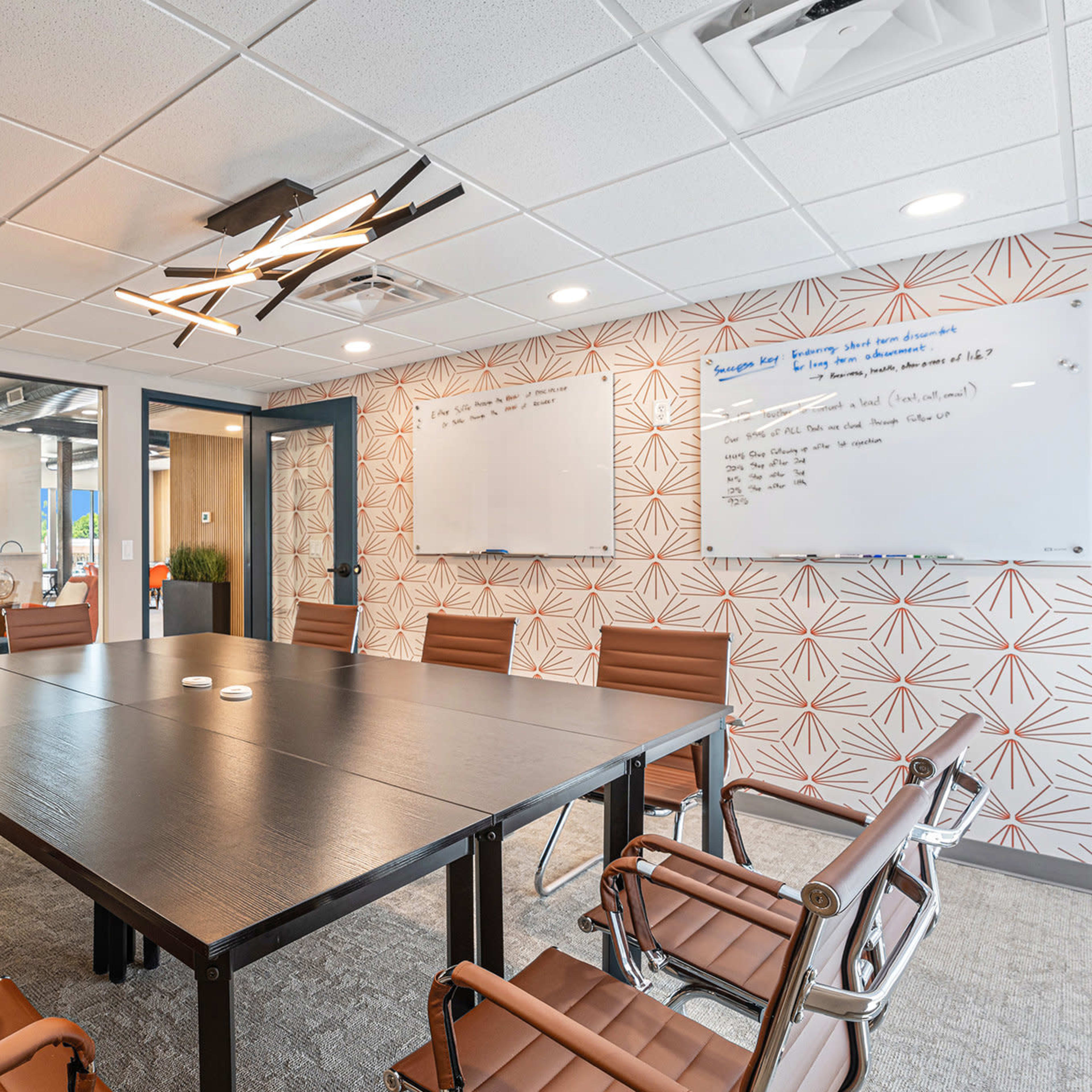 A modern conference room with a large rectangular table surrounded by brown leather chairs, a whiteboard on the wall, and a patterned accent wall.