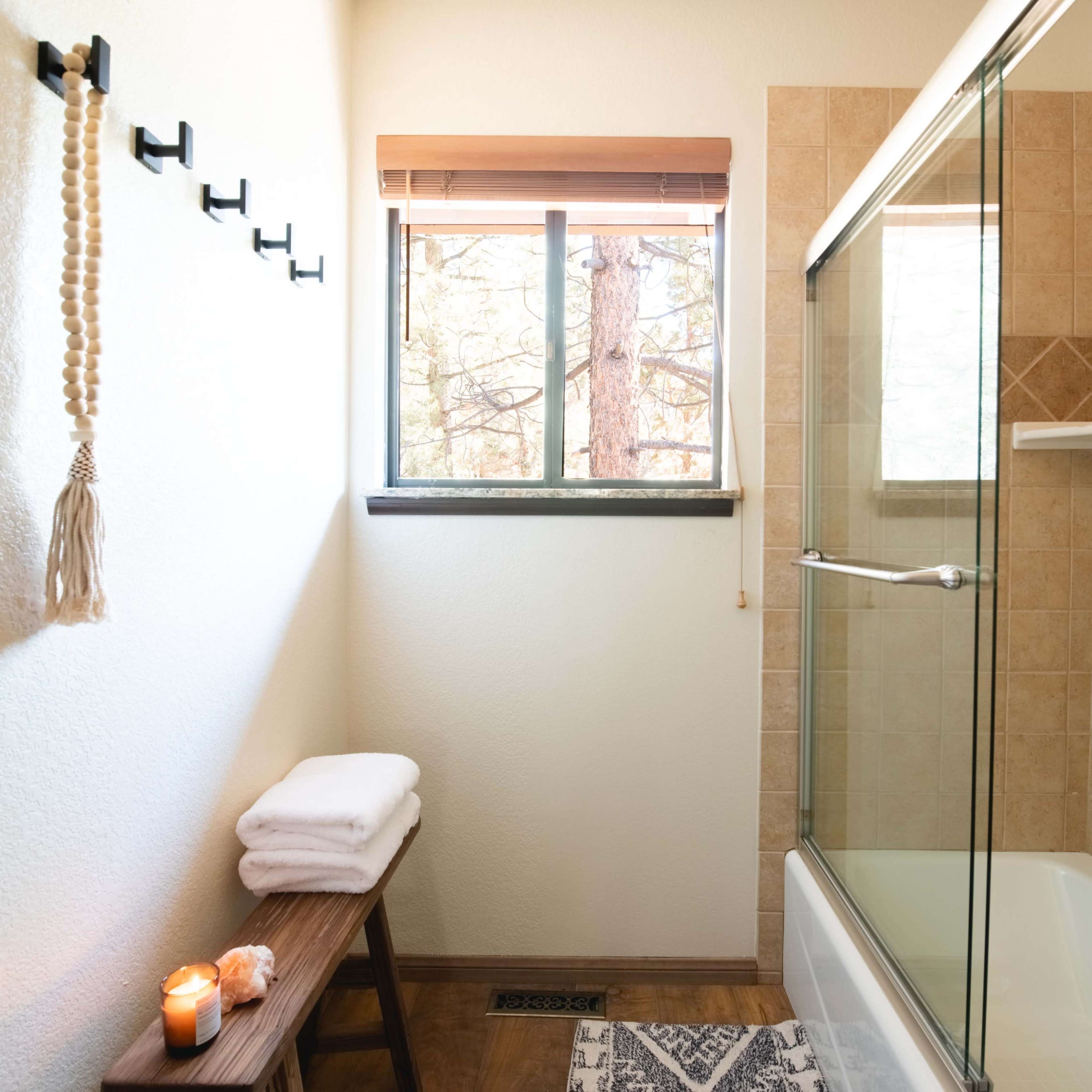 A bathroom with a shower stall, a towel rack, a wooden bench, and a window with a view of trees.