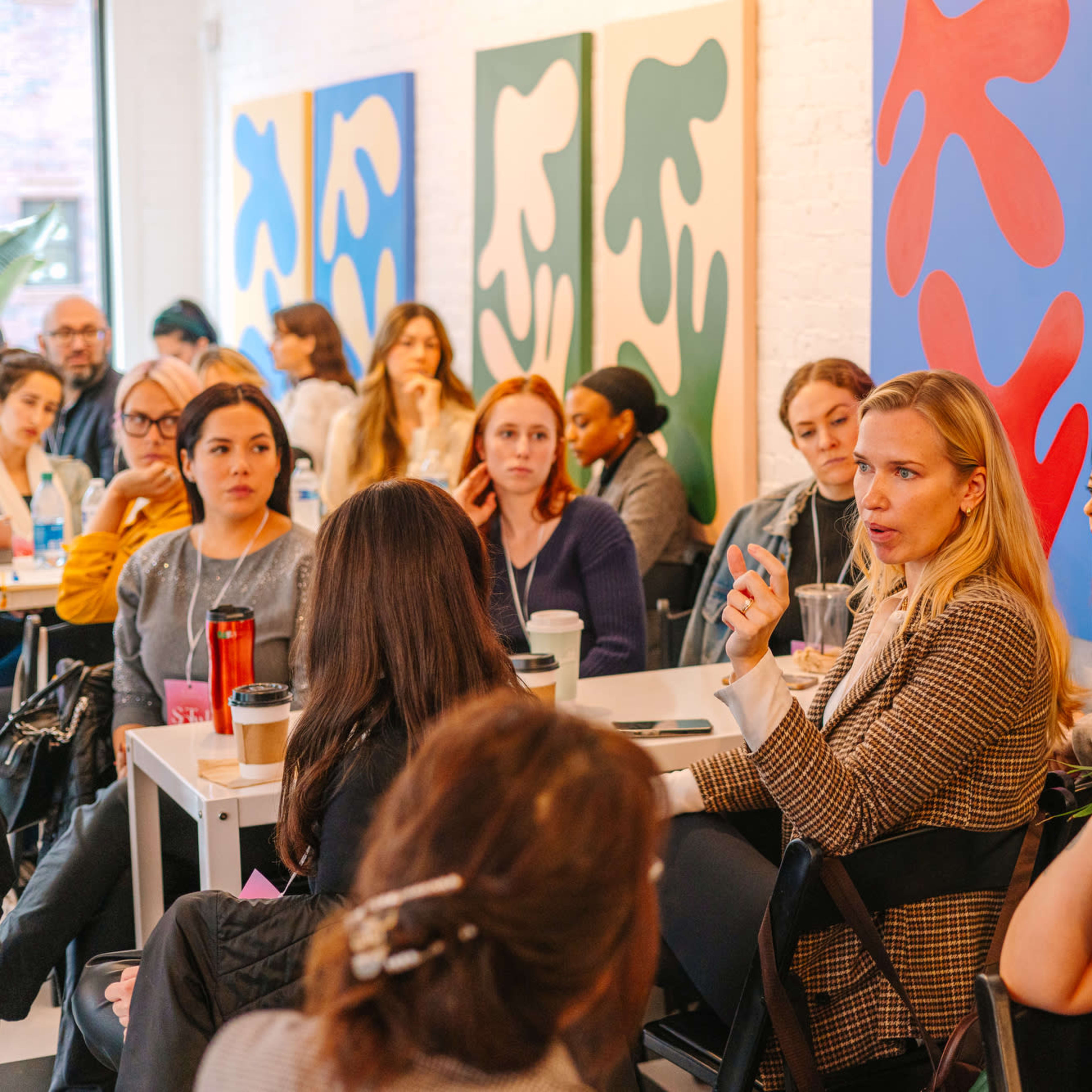 A group of people is seated in a bright room with large colorful abstract paintings on the walls, engaged in a discussion around a table.