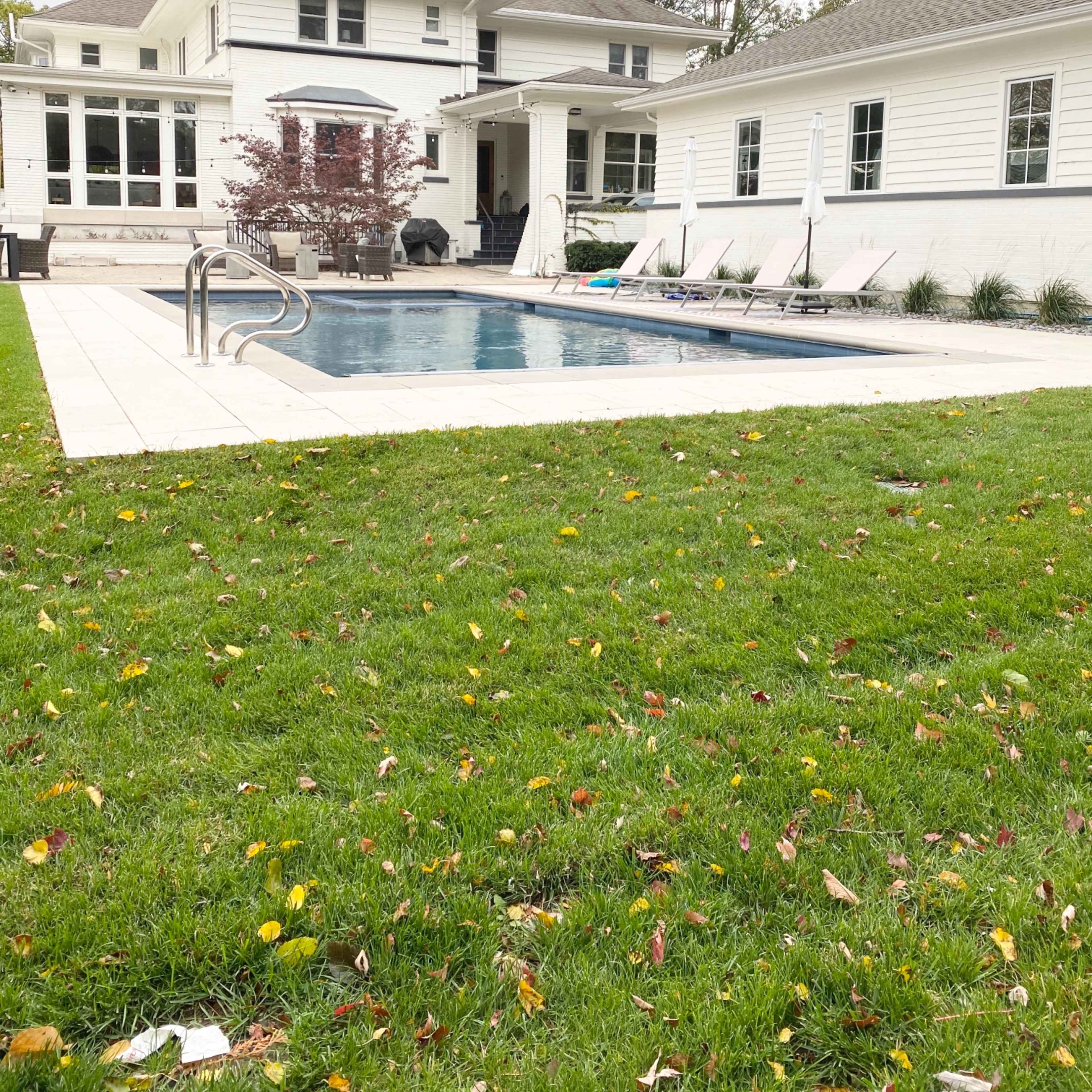 A rectangular swimming pool surrounded by lounge chairs, situated beside a white house with large windows and a well-maintained lawn.