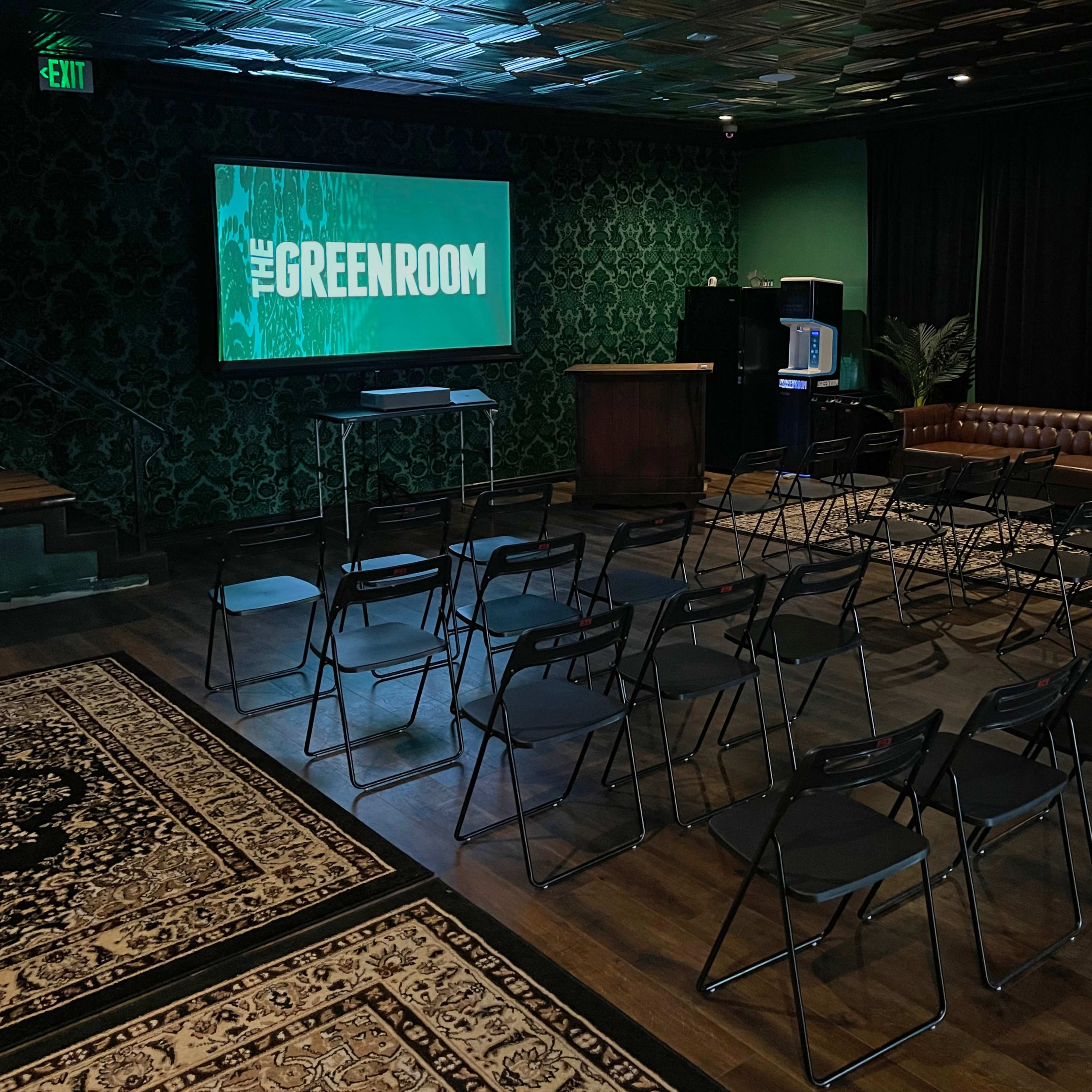 A small event space called "The Greenroom," arranged with rows of black chairs facing a screen, and decorated with a dark green and patterned wall.