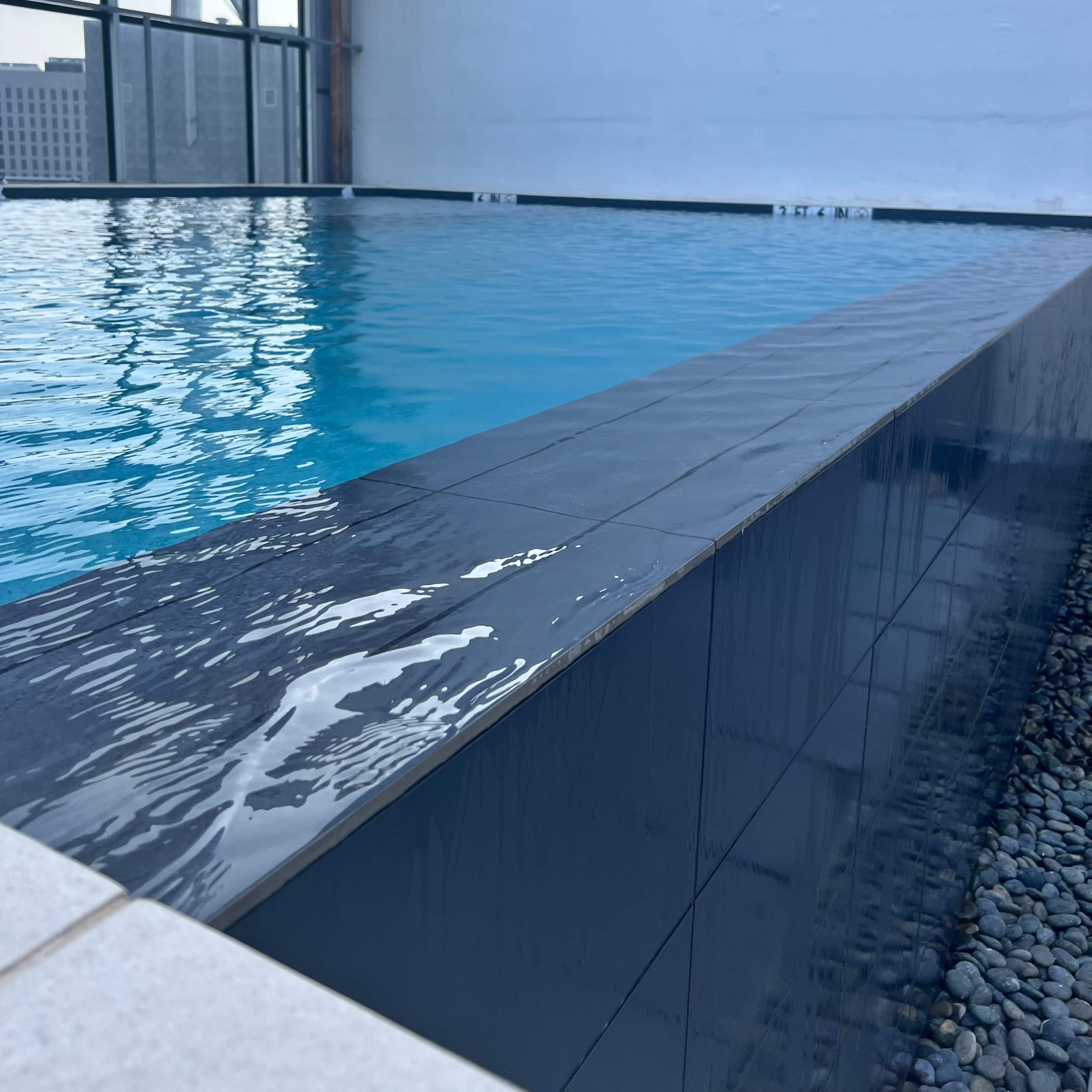 A rooftop swimming pool with a sleek dark edge reflecting the water's surface, situated next to a glass wall.