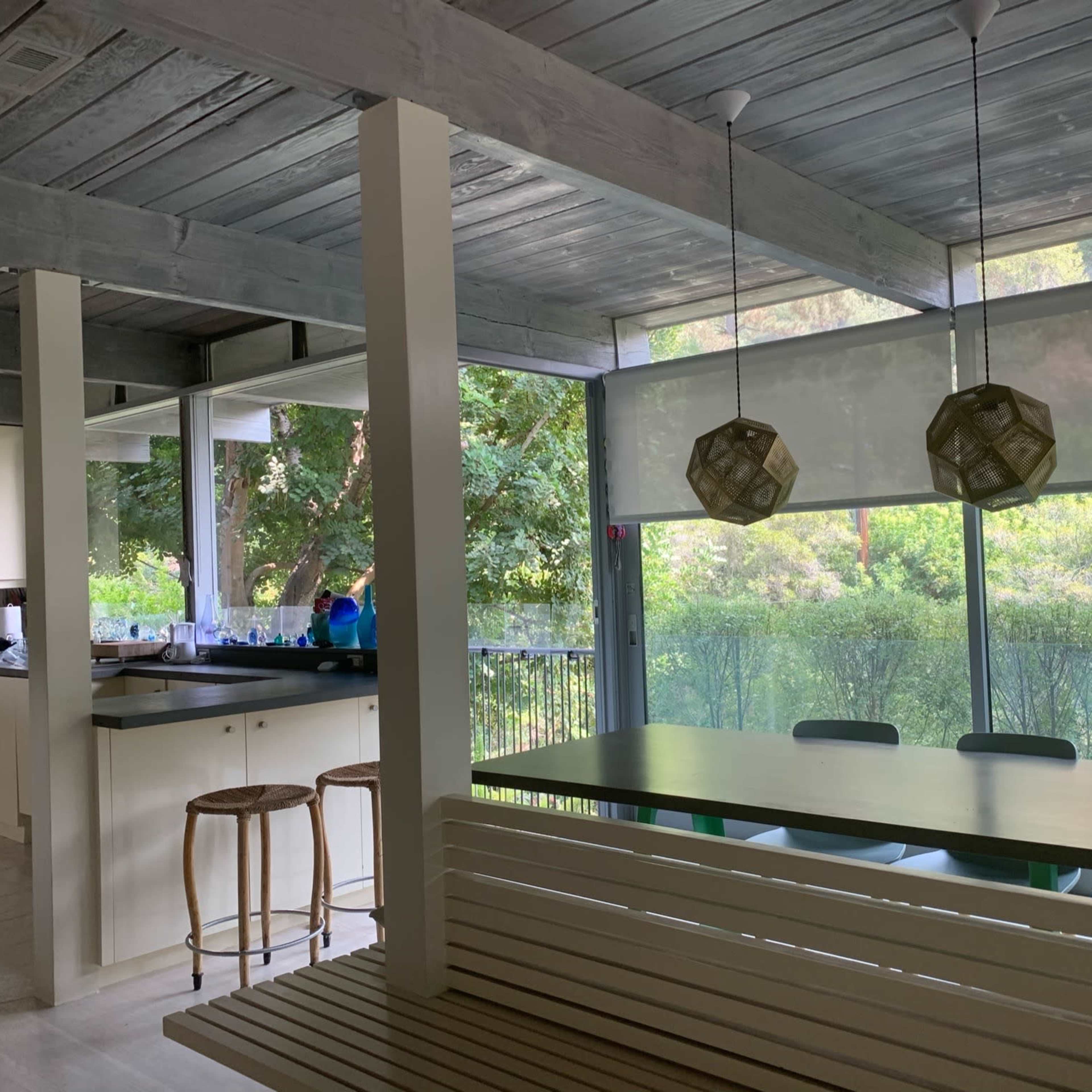 A modern kitchen features a large dining table and glass walls overlooking a lush outdoor area, with pendant lights hanging above.