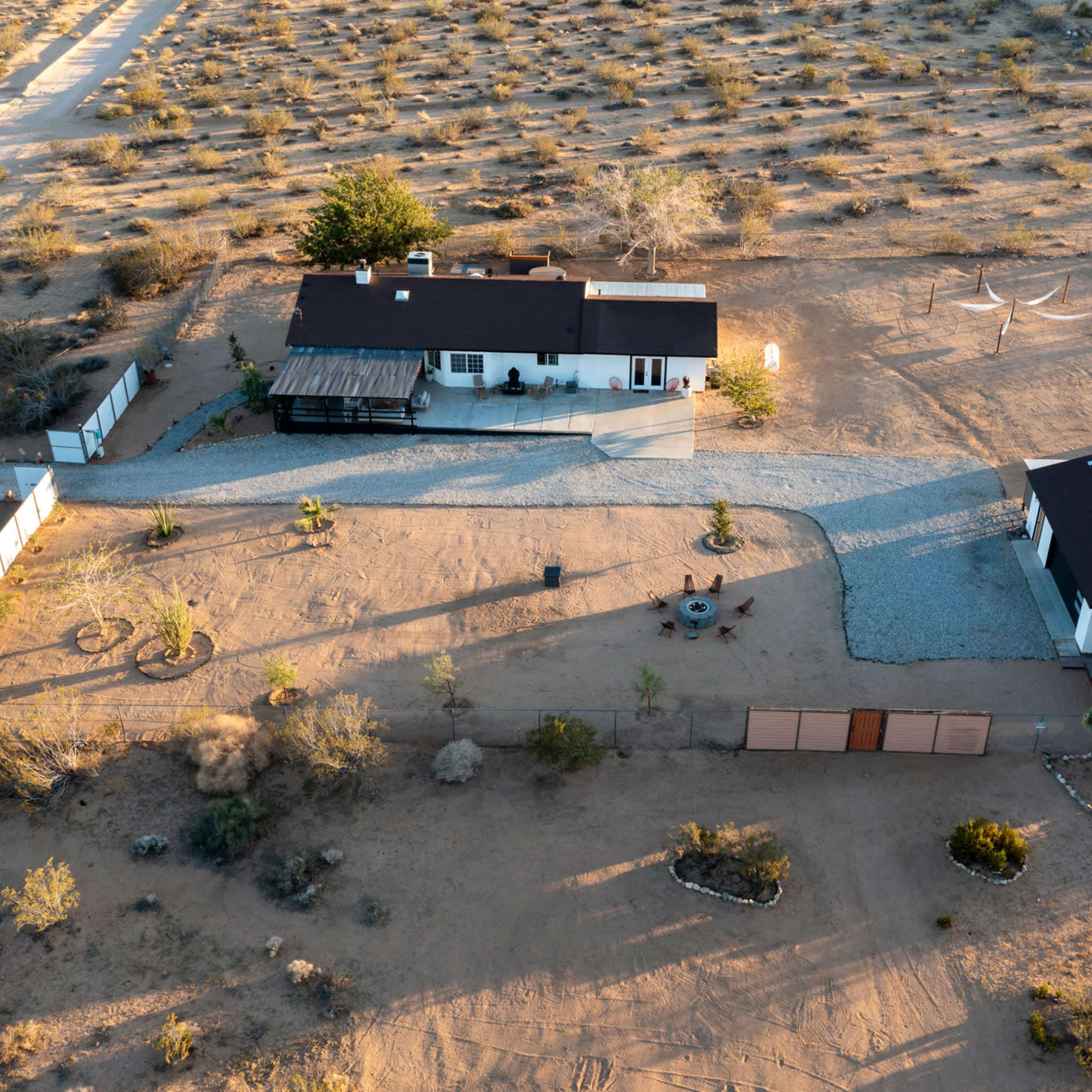 An aerial view of a desert property featuring a main house and a smaller adjacent building, surrounded by sparse vegetation and gravel pathways.