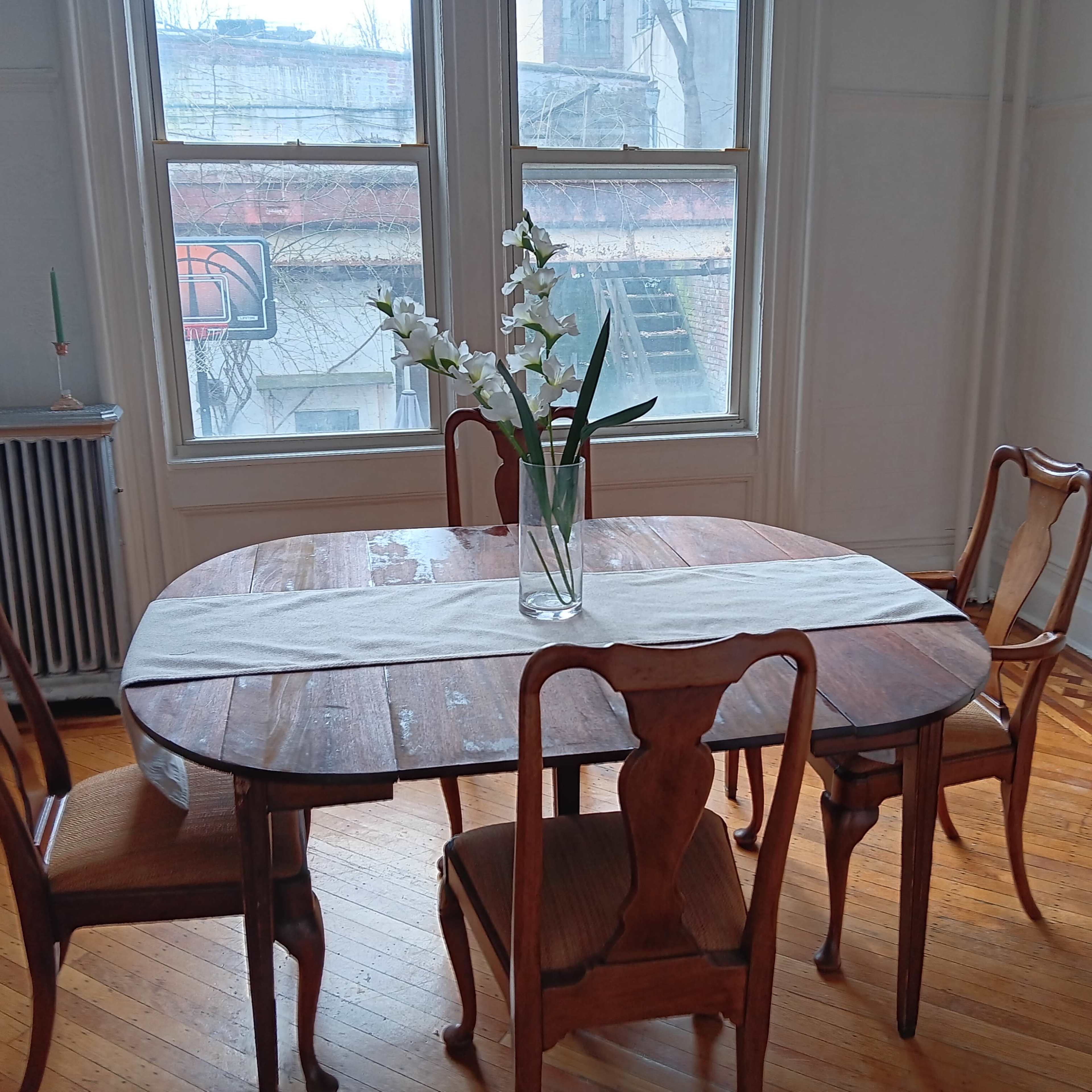 A wooden dining table with four chairs is set near two large windows, displaying a vase of flowers on the table and a plant on the windowsill.
