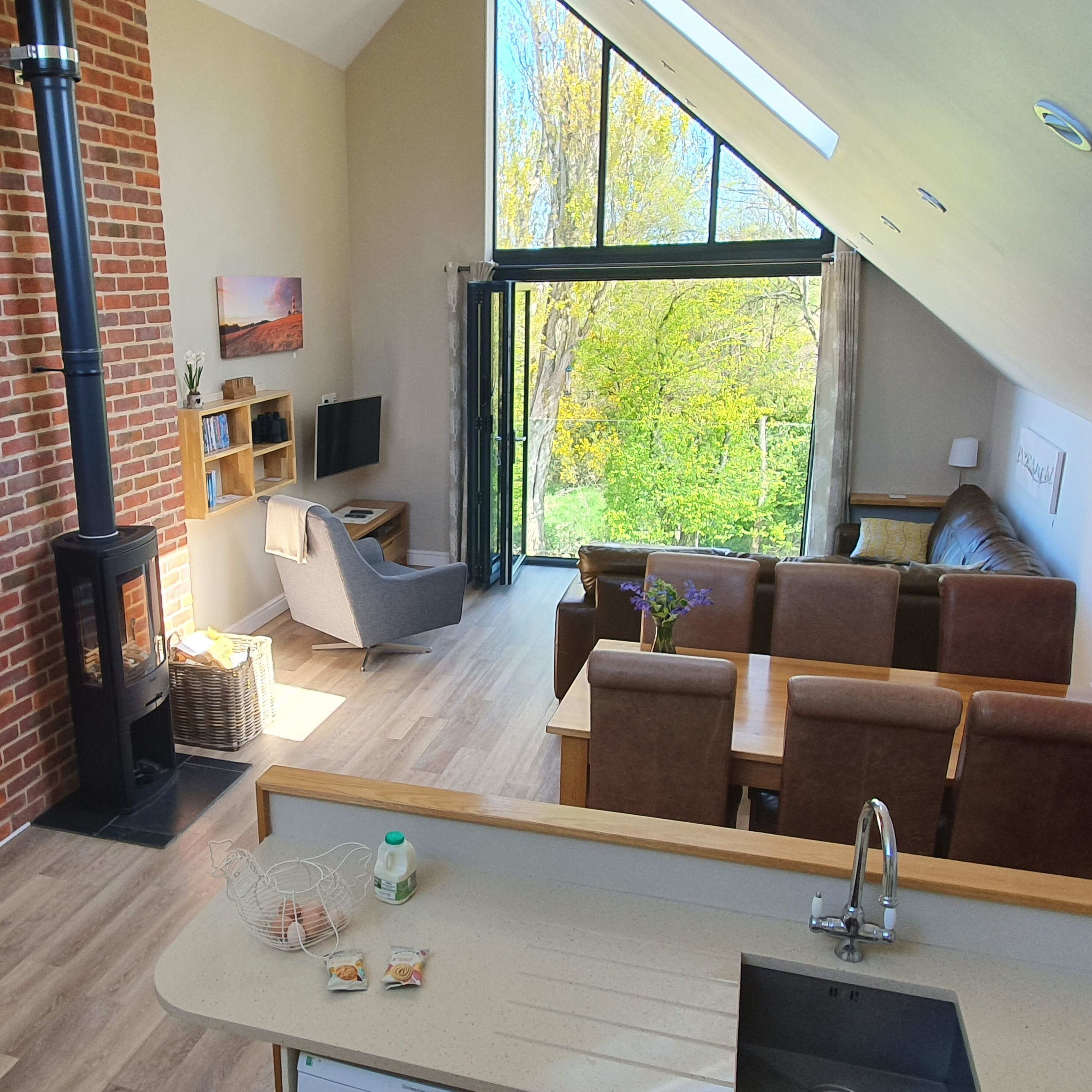 A modern open-concept living area with a kitchen, dining table, and large windows overlooking a green outdoor space.