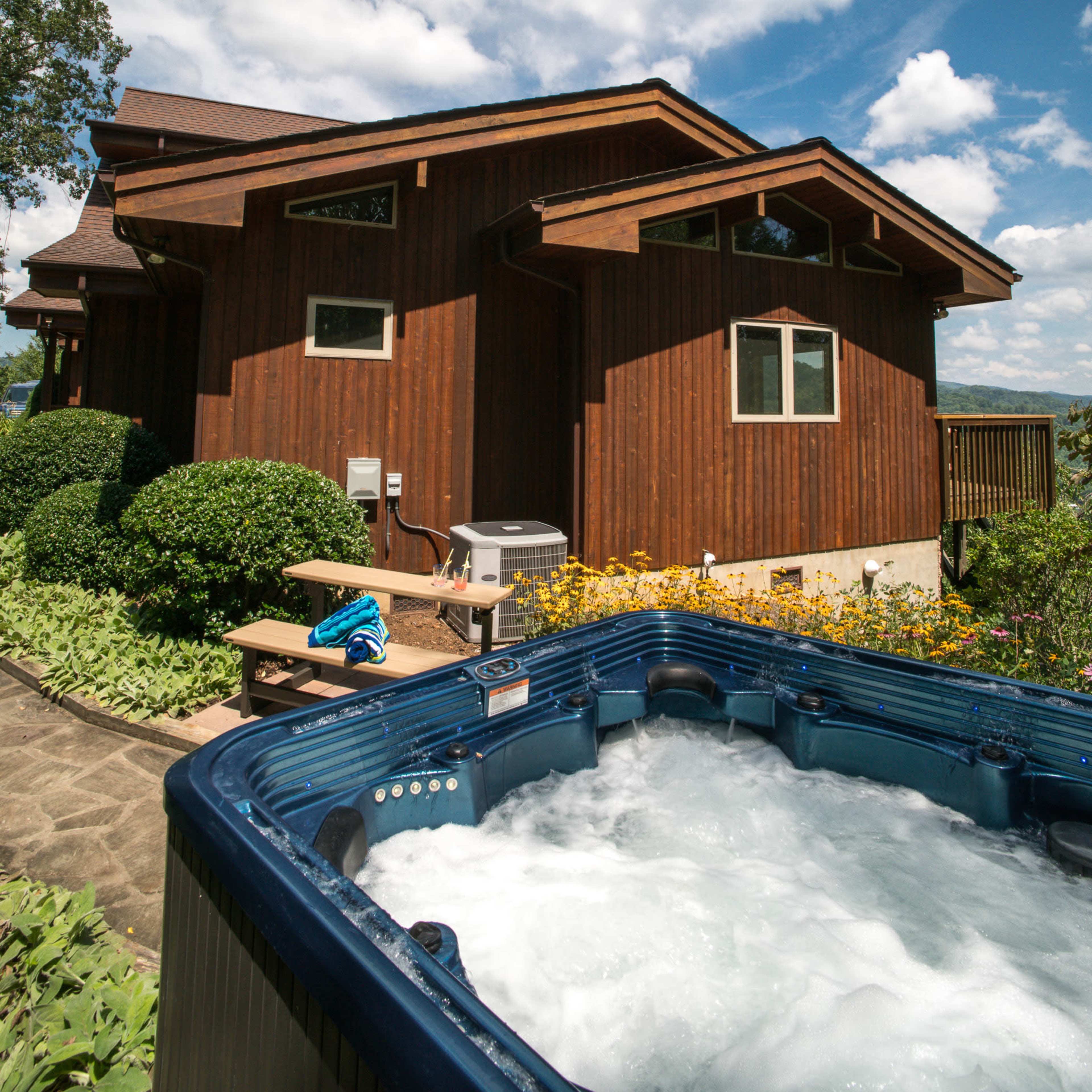 A hot tub bubbles beside a stone pathway leading to a wooden house surrounded by greenery and flowers.