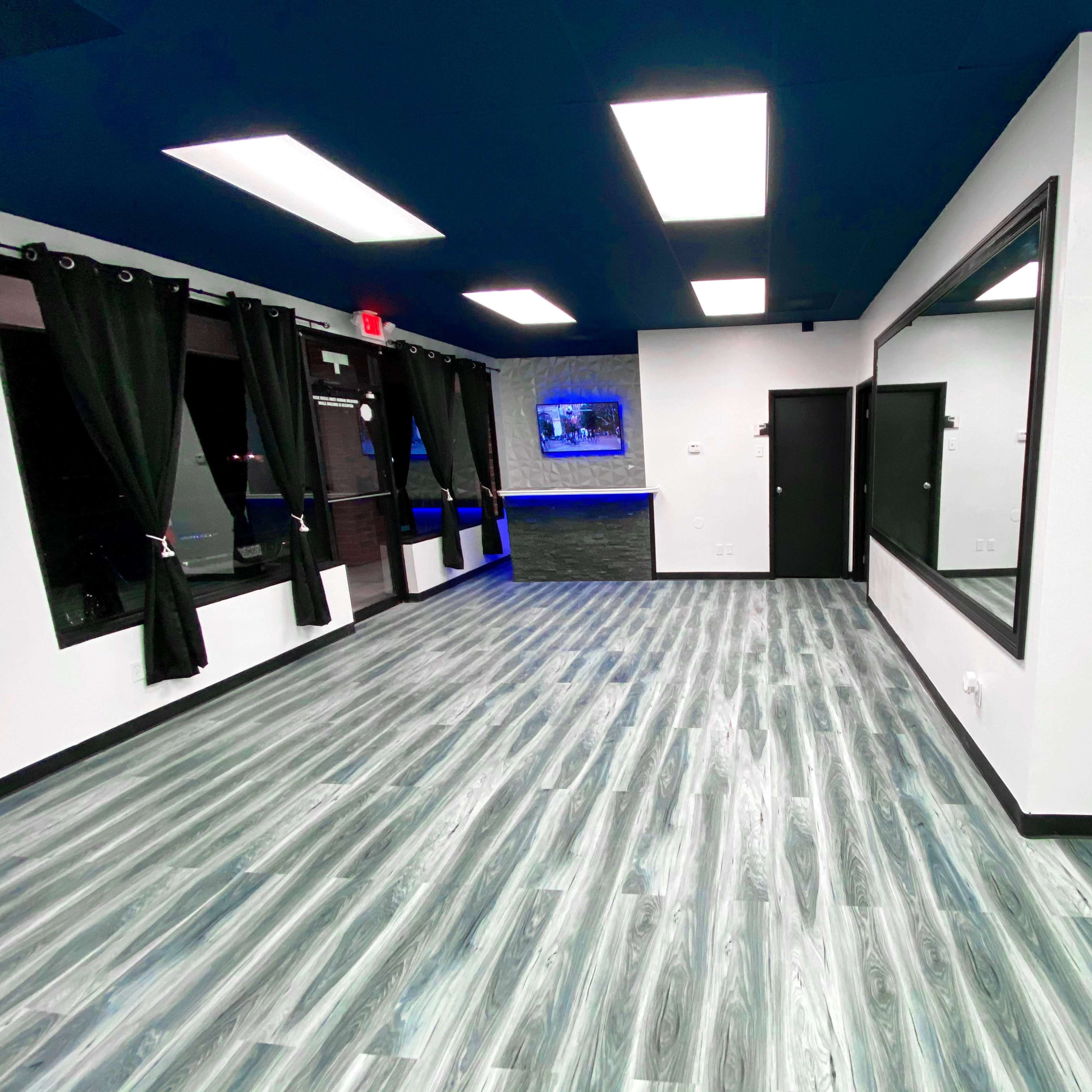 A spacious room with a dark blue ceiling, modern lighting, and a light gray wooden floor, featuring large mirrors and black curtains along the windows.
