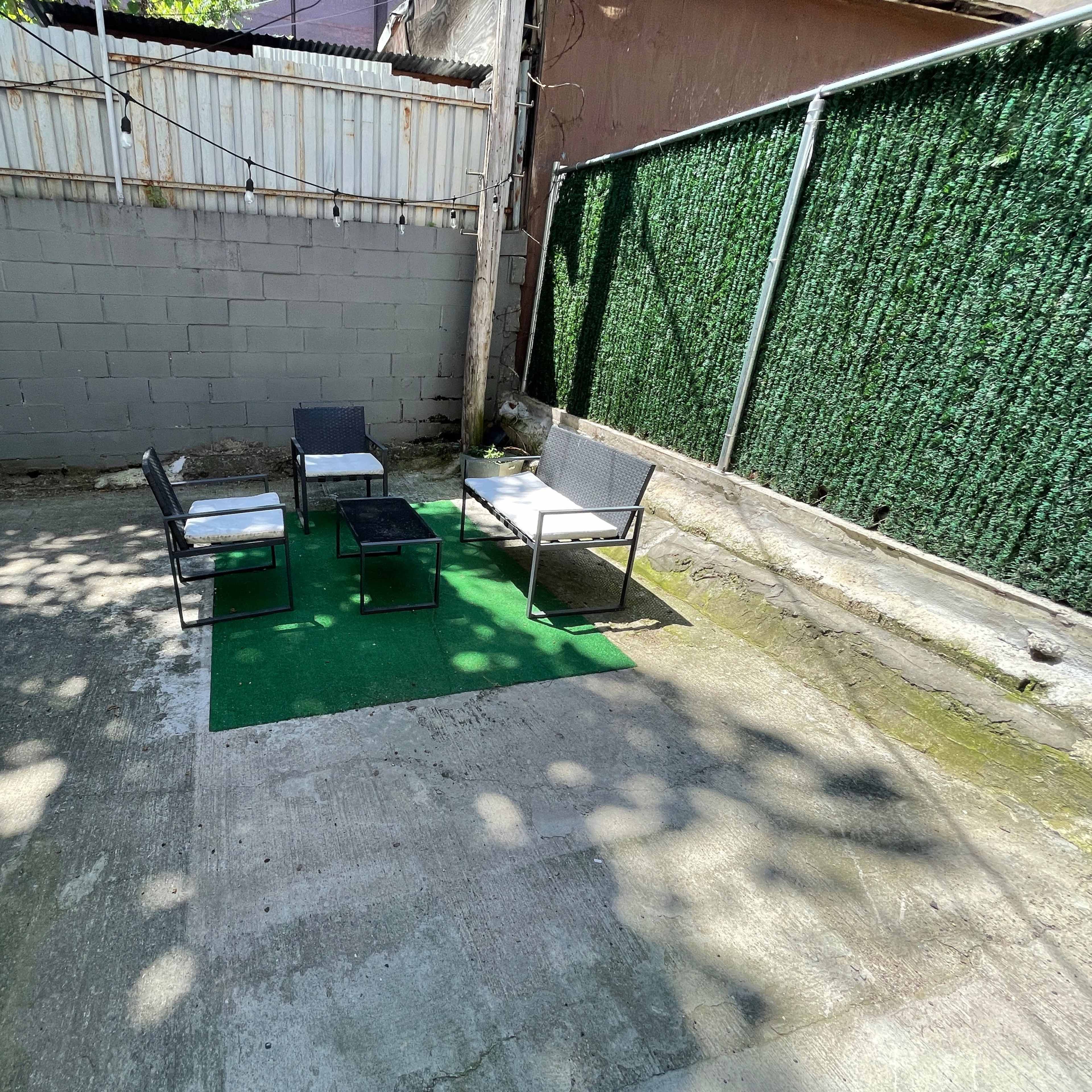 A small, concrete outdoor area featuring four chairs and a table arranged on a green mat, with a wall covered in green foliage in the background.