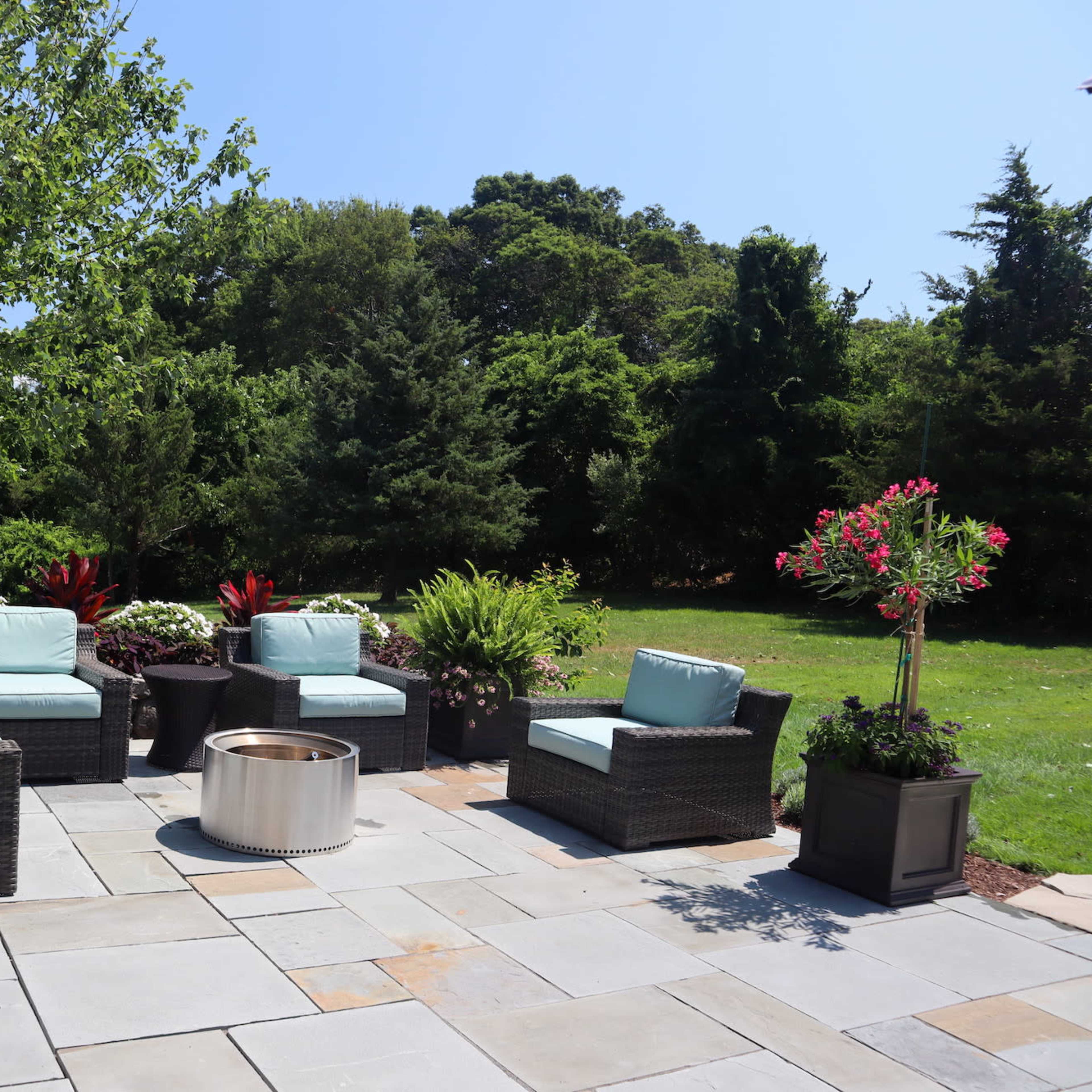 A patio area features four wicker chairs with light blue cushions arranged around a circular fire pit, surrounded by green shrubs and decorative planters.