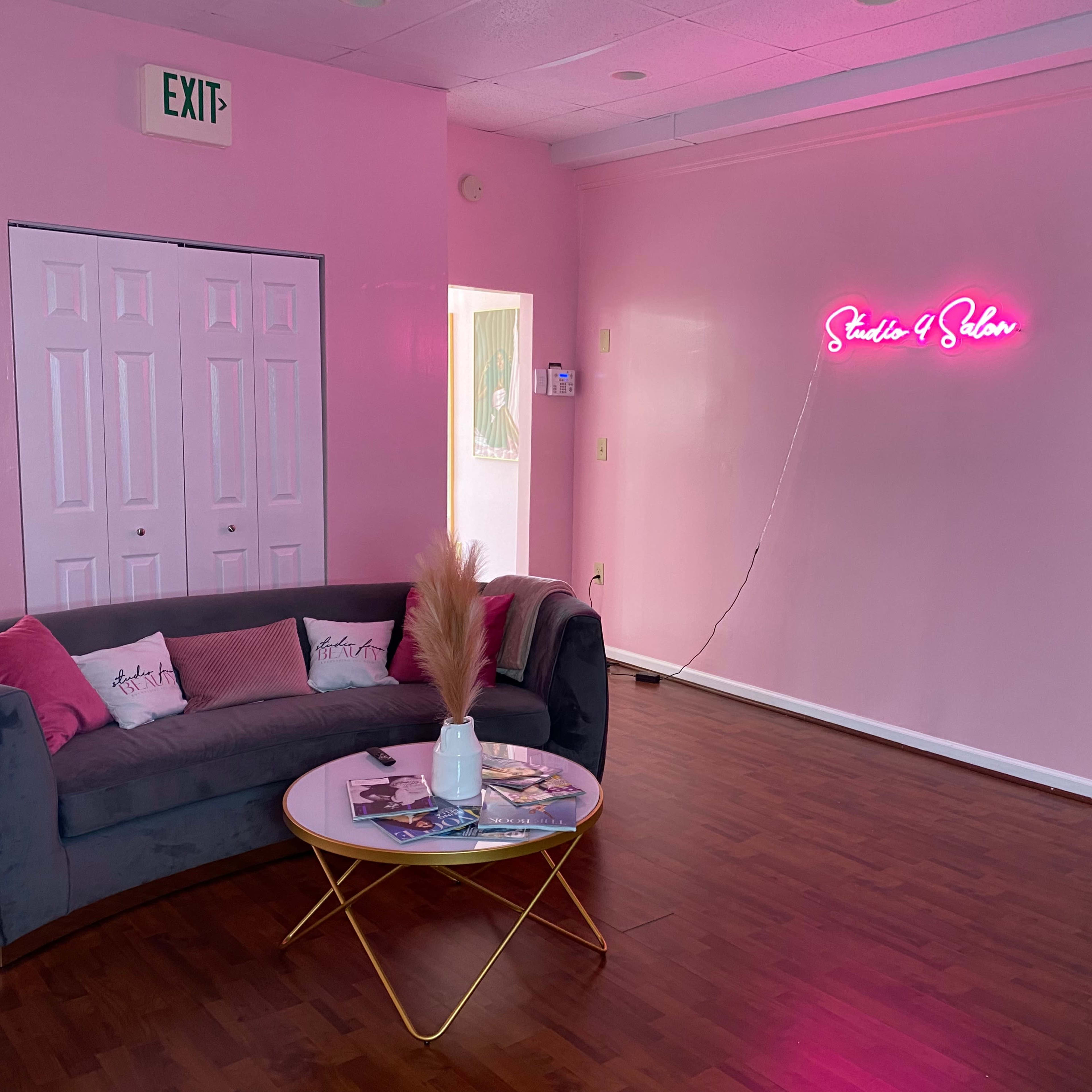 A pink-walled room features a gray couch with decorative pillows, a round coffee table, and a neon sign that reads "Studio & Salon."