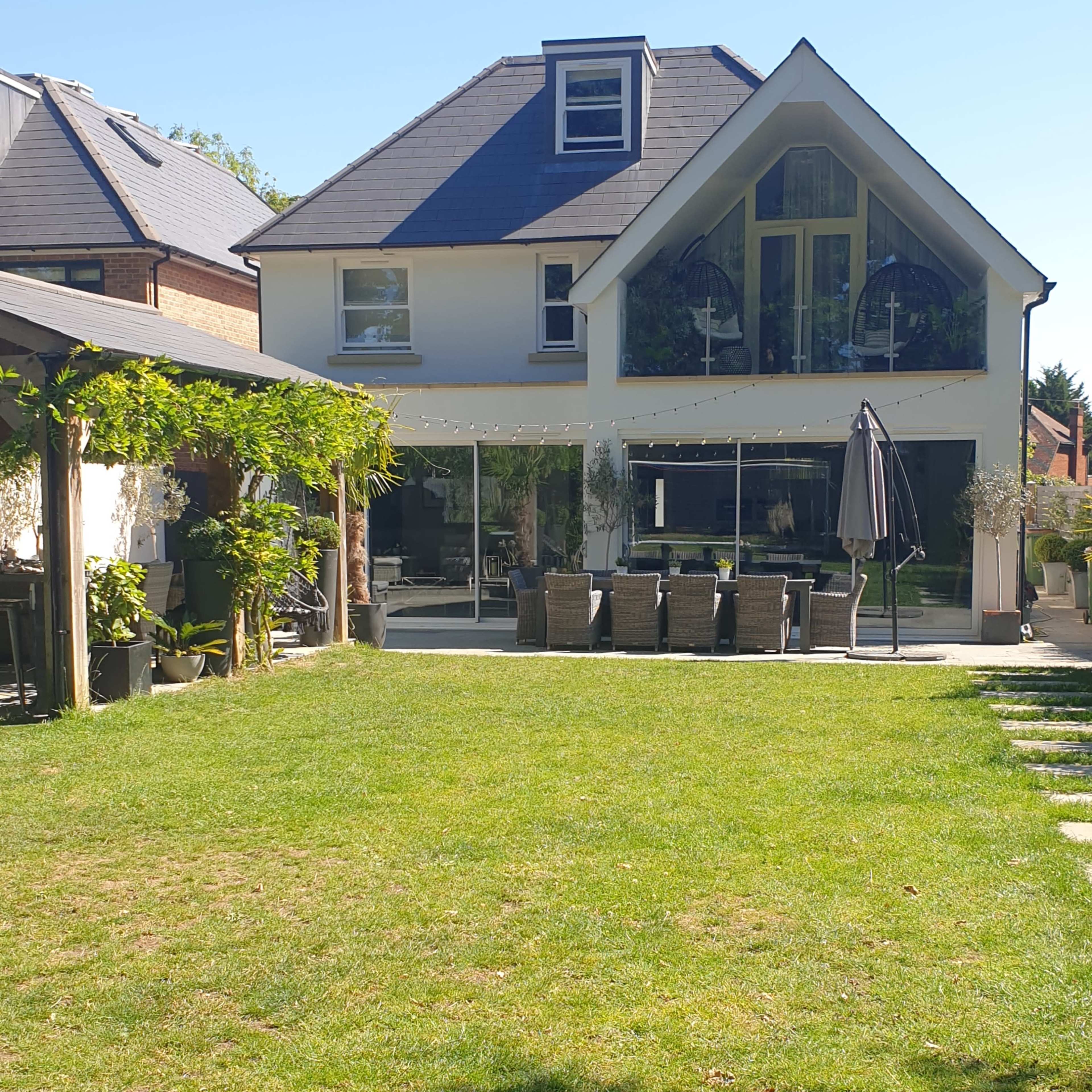 A modern two-story house with a large glass patio overlooks a well-maintained green lawn and patio seating area.