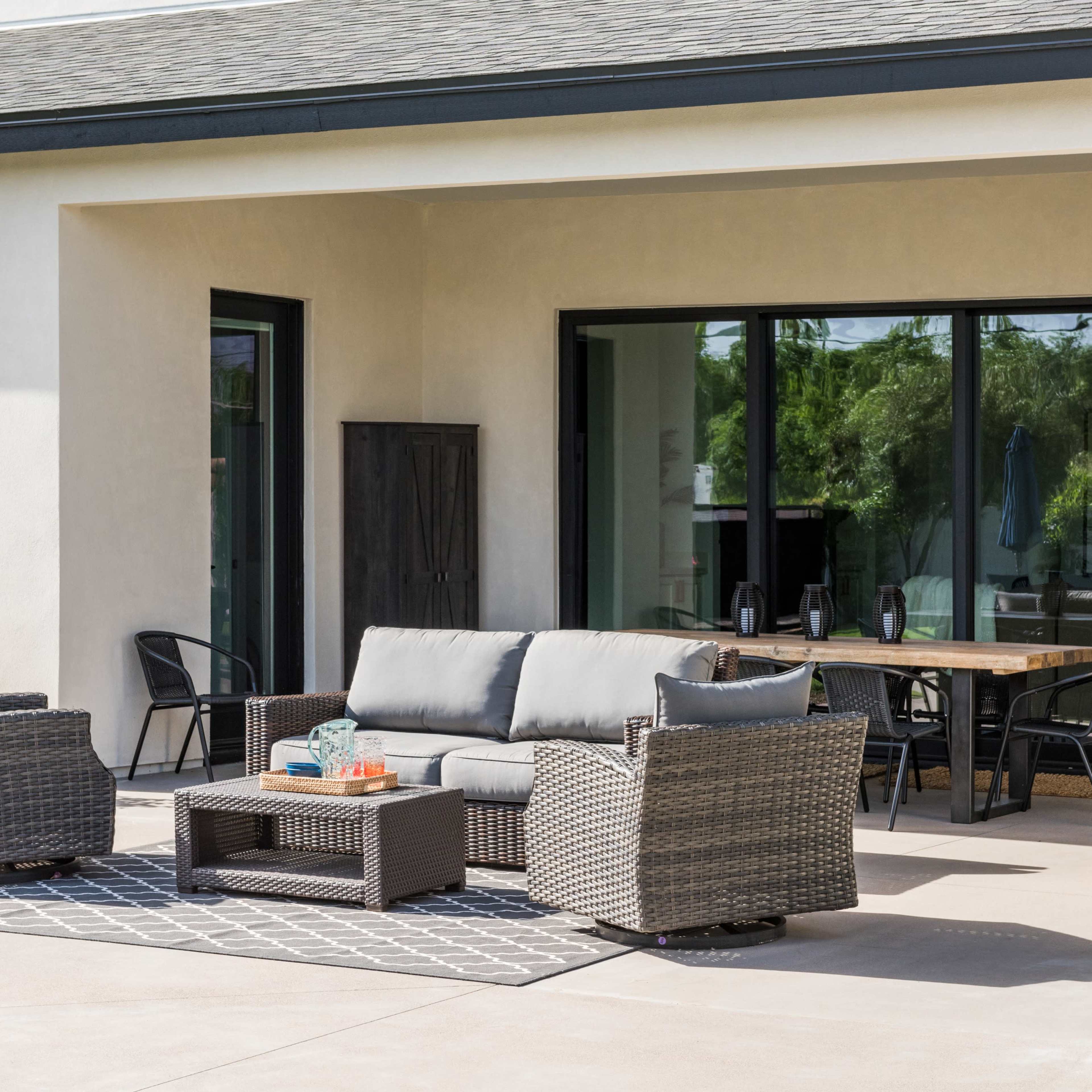 A modern patio features a gray sofa, two matching chairs, and a coffee table on a patterned rug, surrounded by sliding glass doors and a dining area.