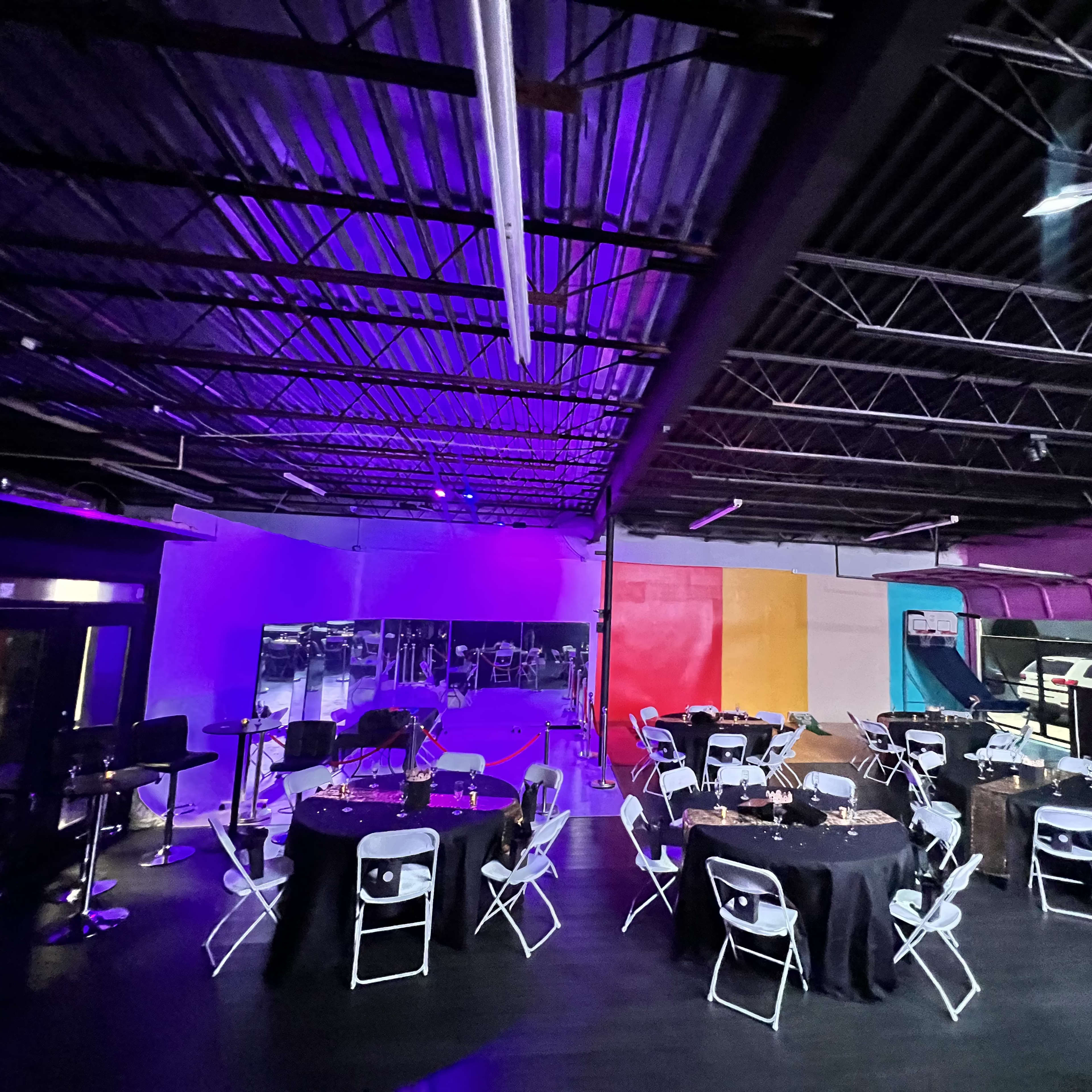 An event space with round tables and chairs set up under purple and blue lighting, featuring a dance floor area in the background.