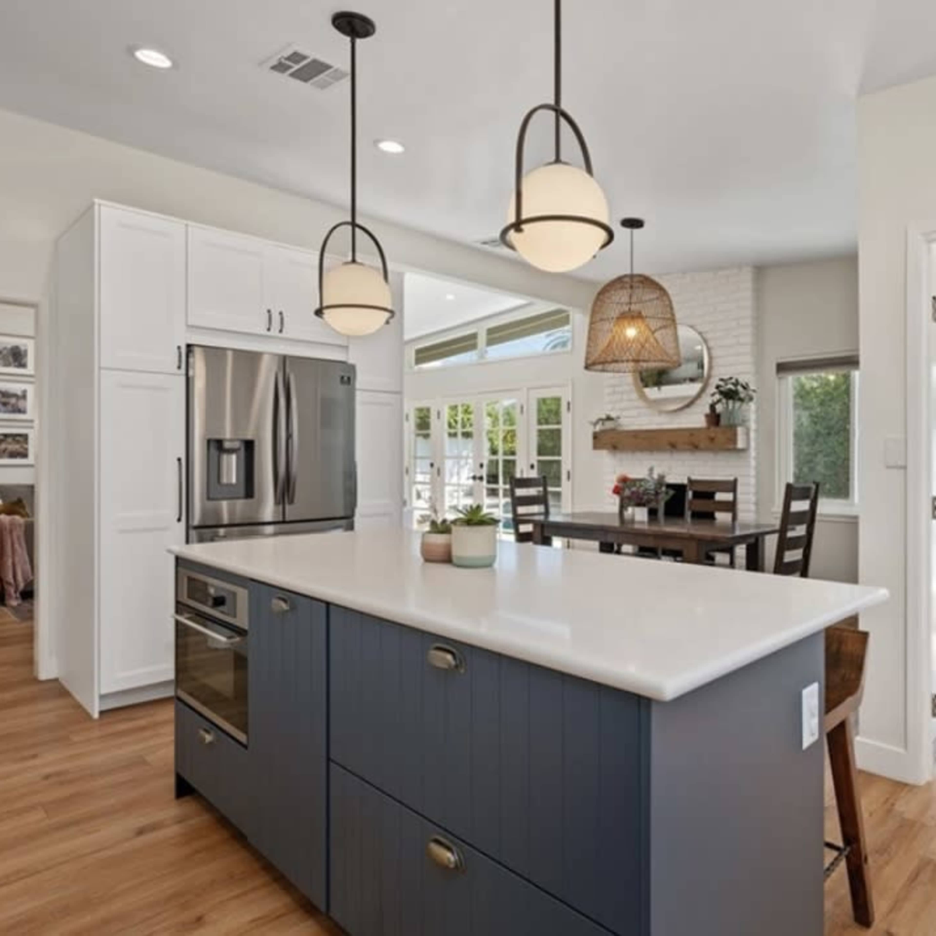 A modern kitchen with a central island featuring a built-in oven and a white countertop, connected to a dining area and a door leading to an outdoor space.