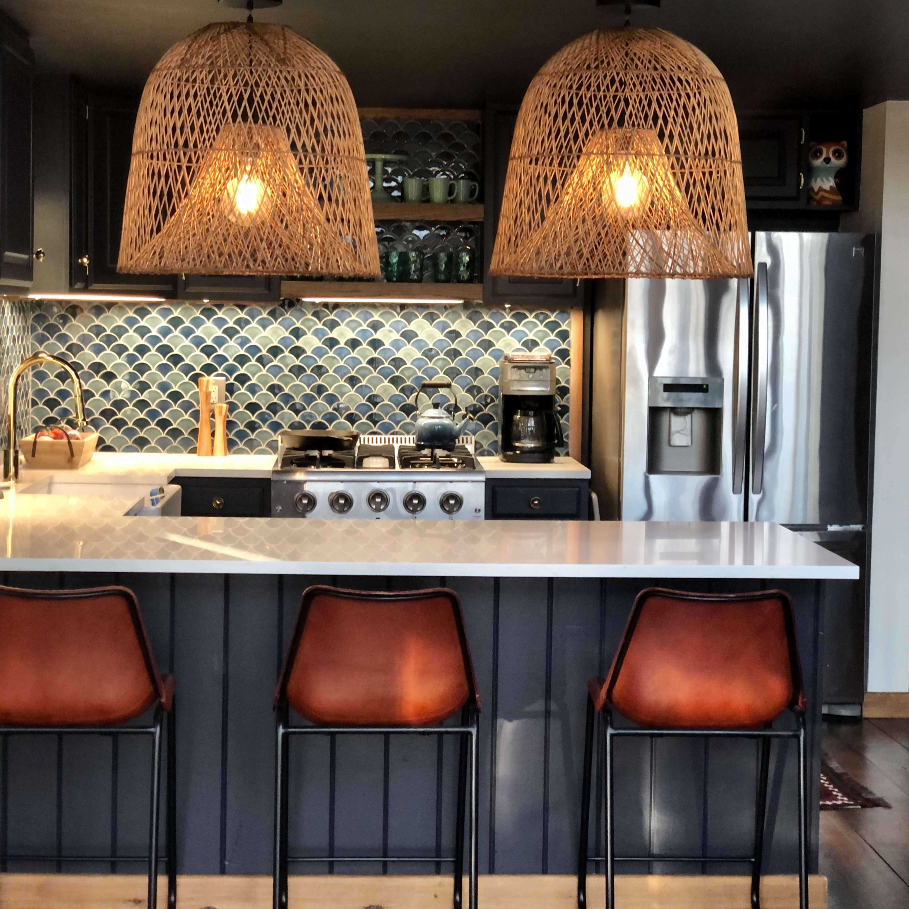 A modern kitchen features a marble island with three black bar stools, two woven pendant lights overhead, and a backdrop of blue tiled walls.