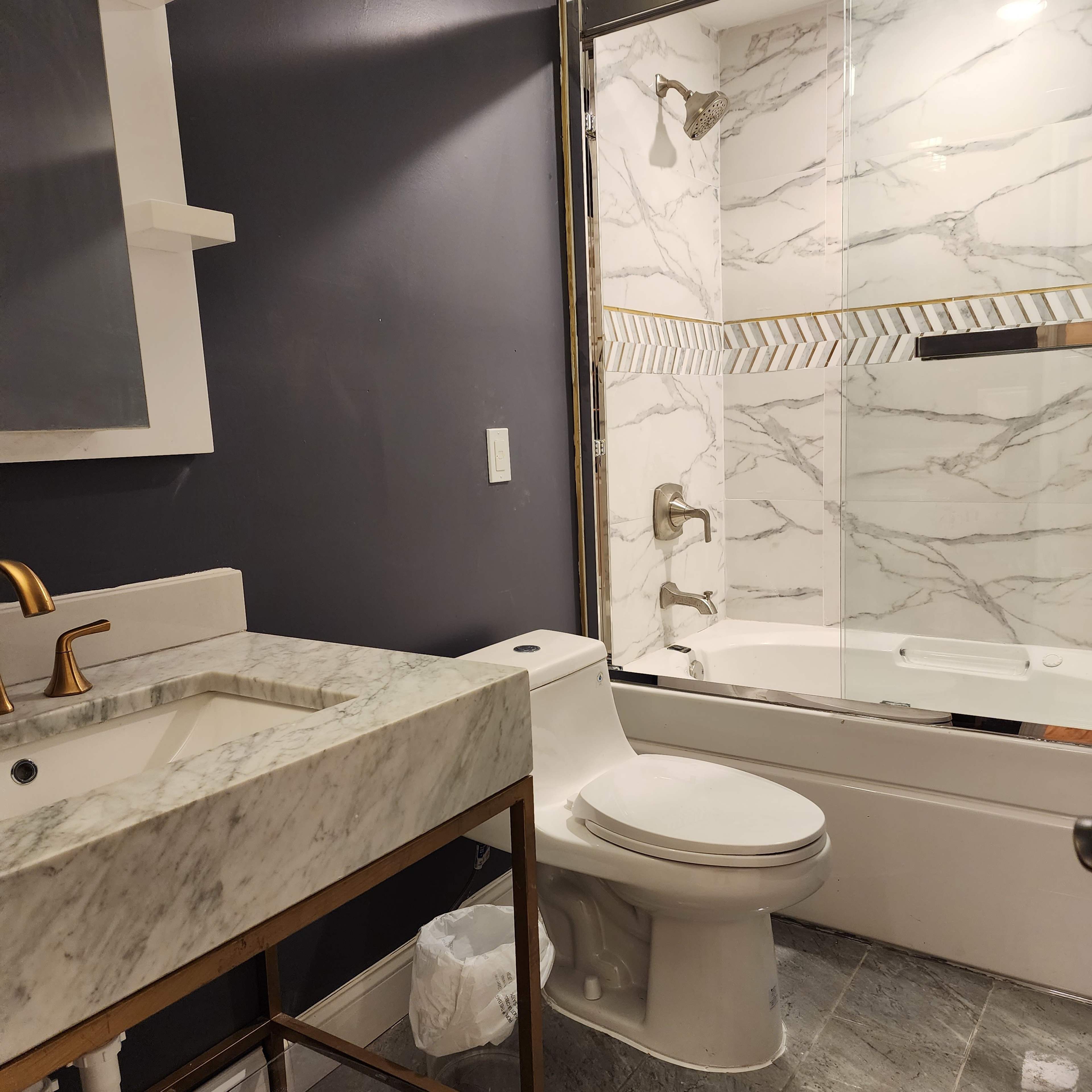 A modern bathroom featuring a marble vanity with a gold faucet, a toilet, and a glass-enclosed shower with marble-patterned tiles.