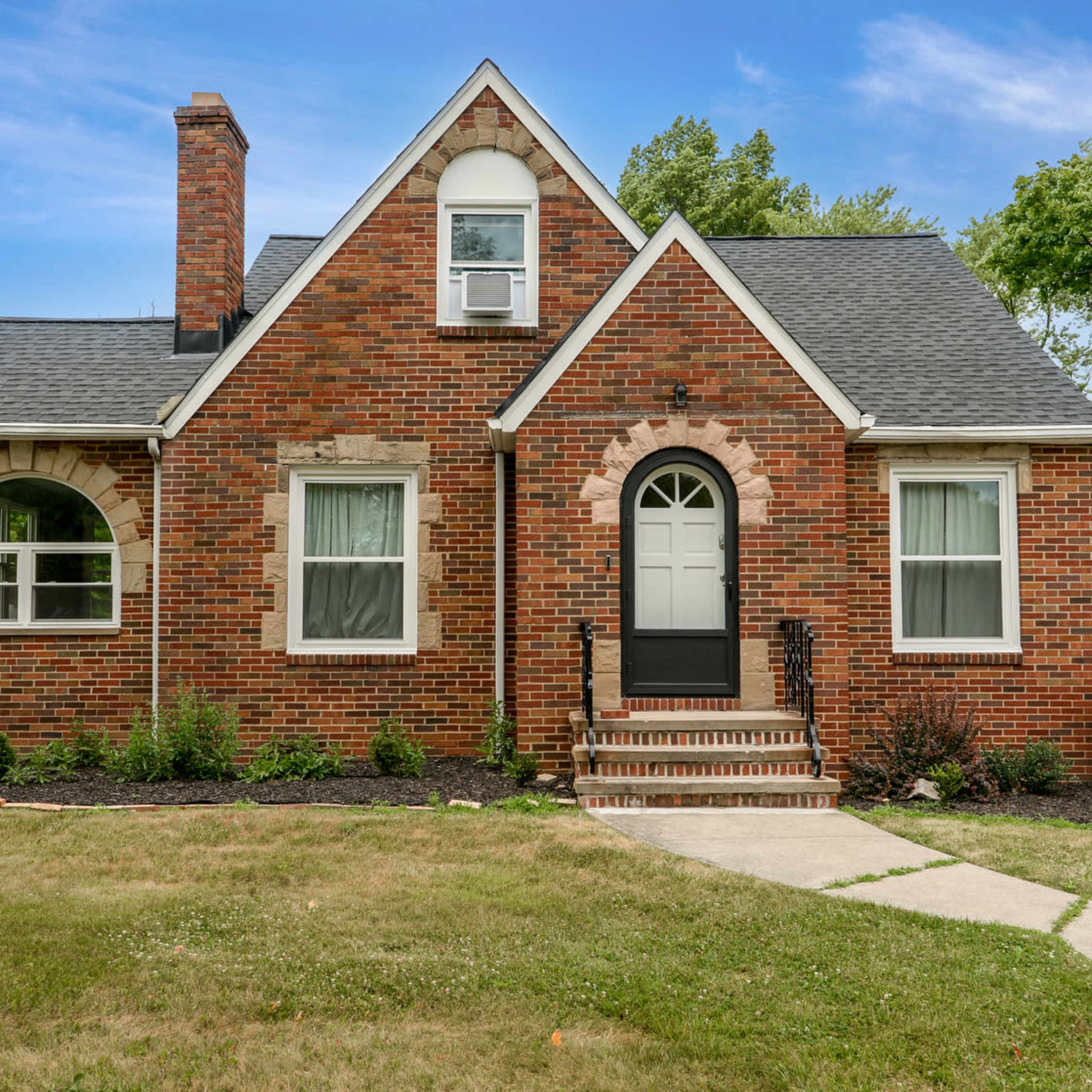 A brick house with a sloped roof, front steps, and a chimney is framed by trees and a well-maintained lawn.