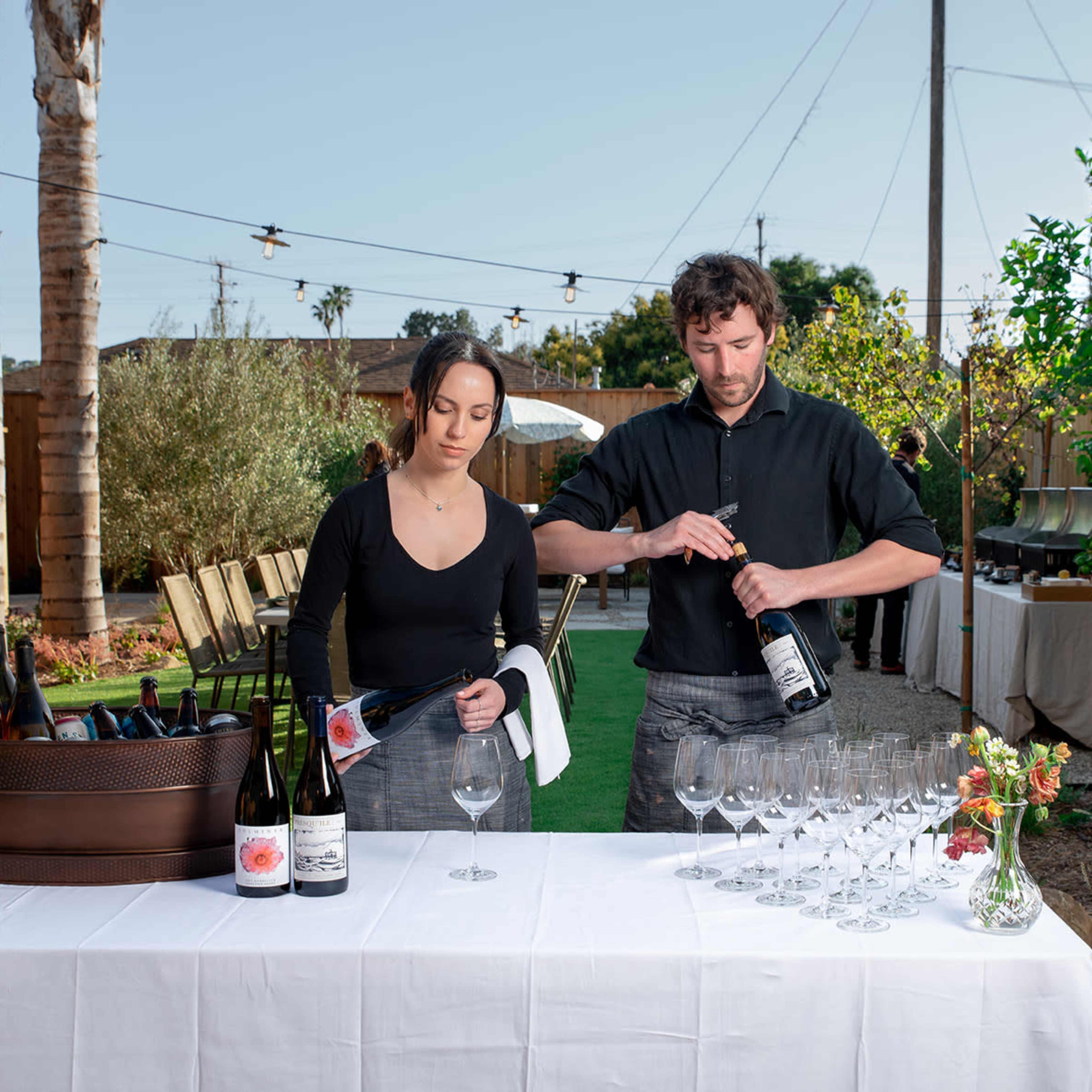 A woman and a man are preparing glasses and pouring wine at an outdoor event with a garden setup.
