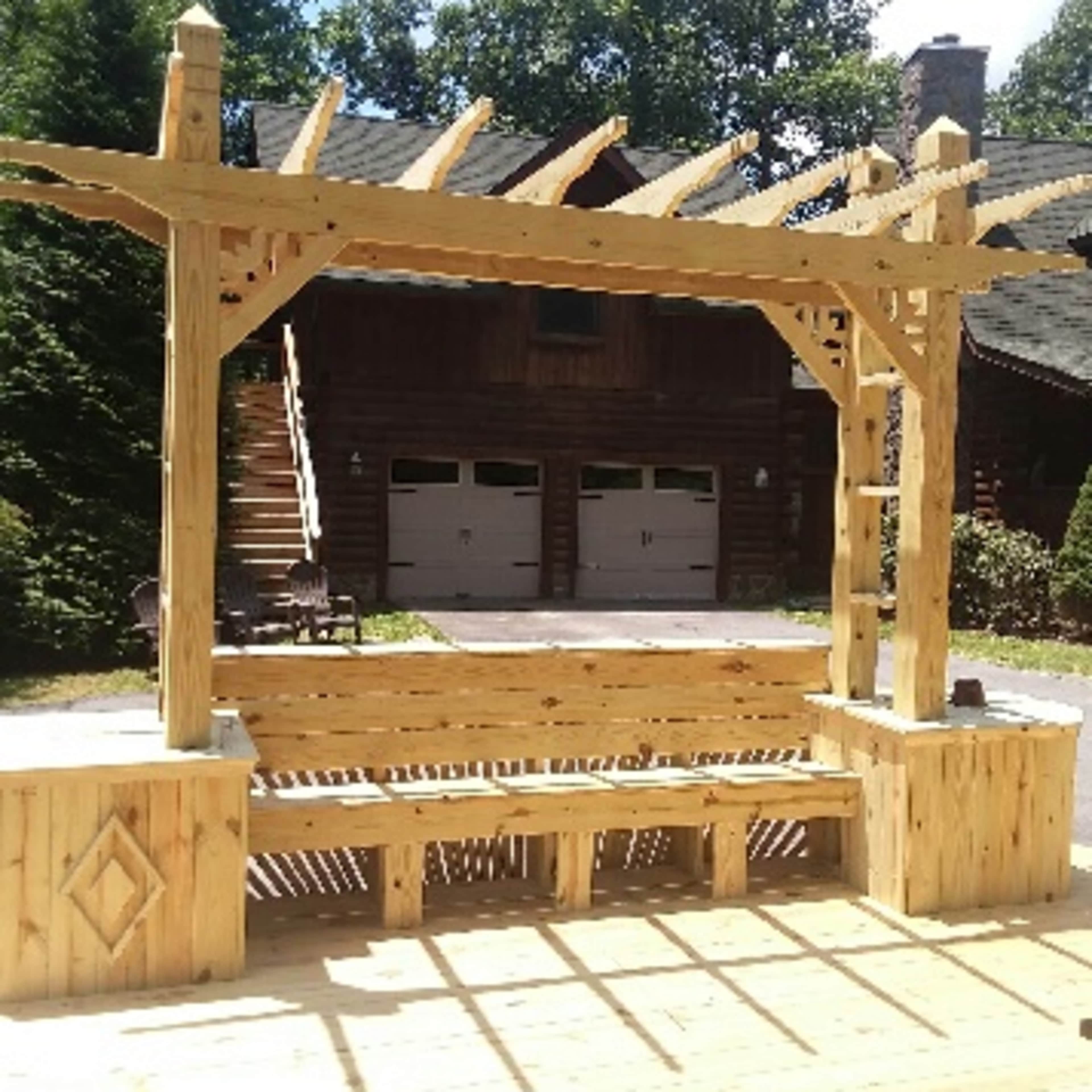 A wooden pergola stands in front of a two-story log cabin, with a deck beneath it and a tree line in the background.