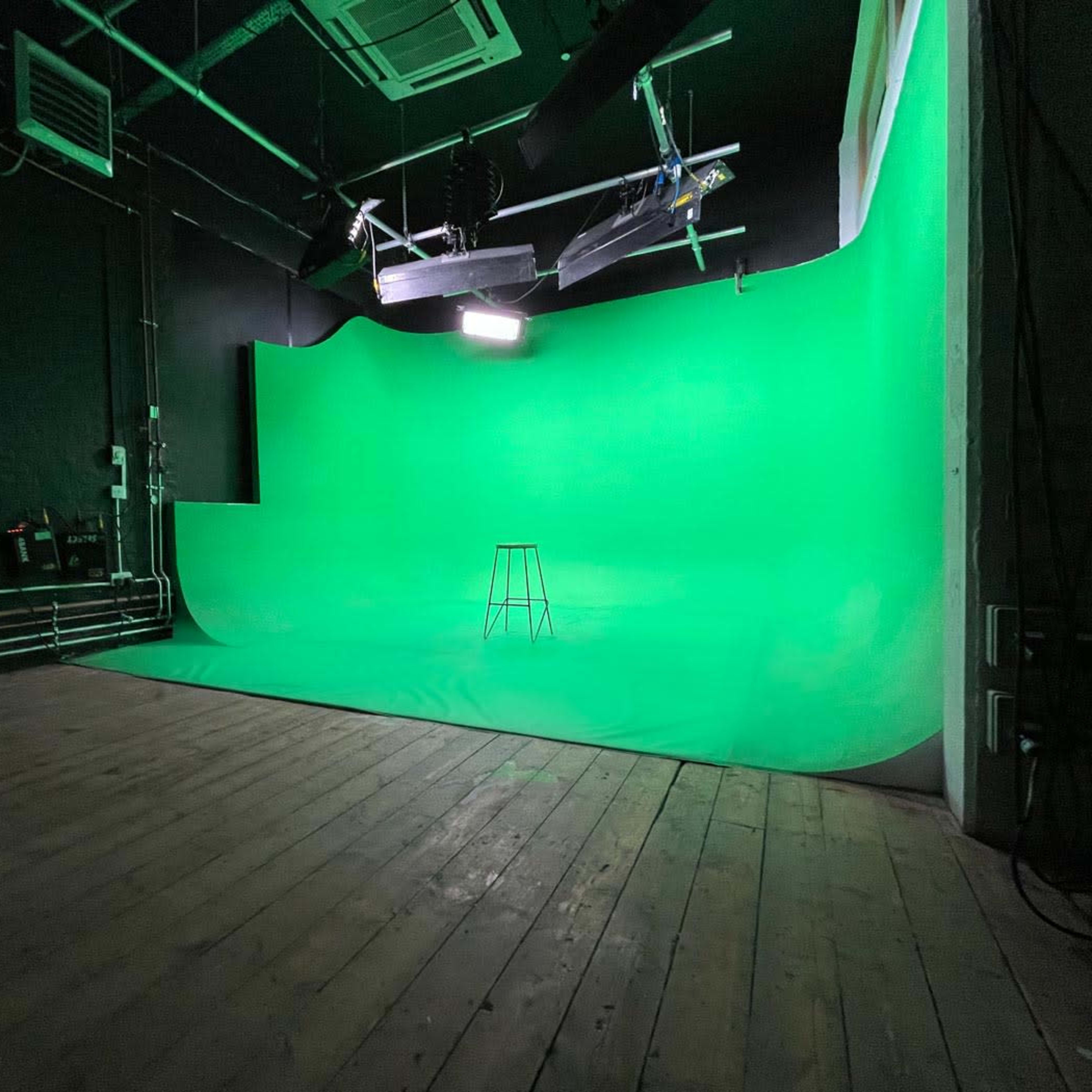 A green screen studio with a bare wooden floor and a single stool positioned in front of a large green backdrop.