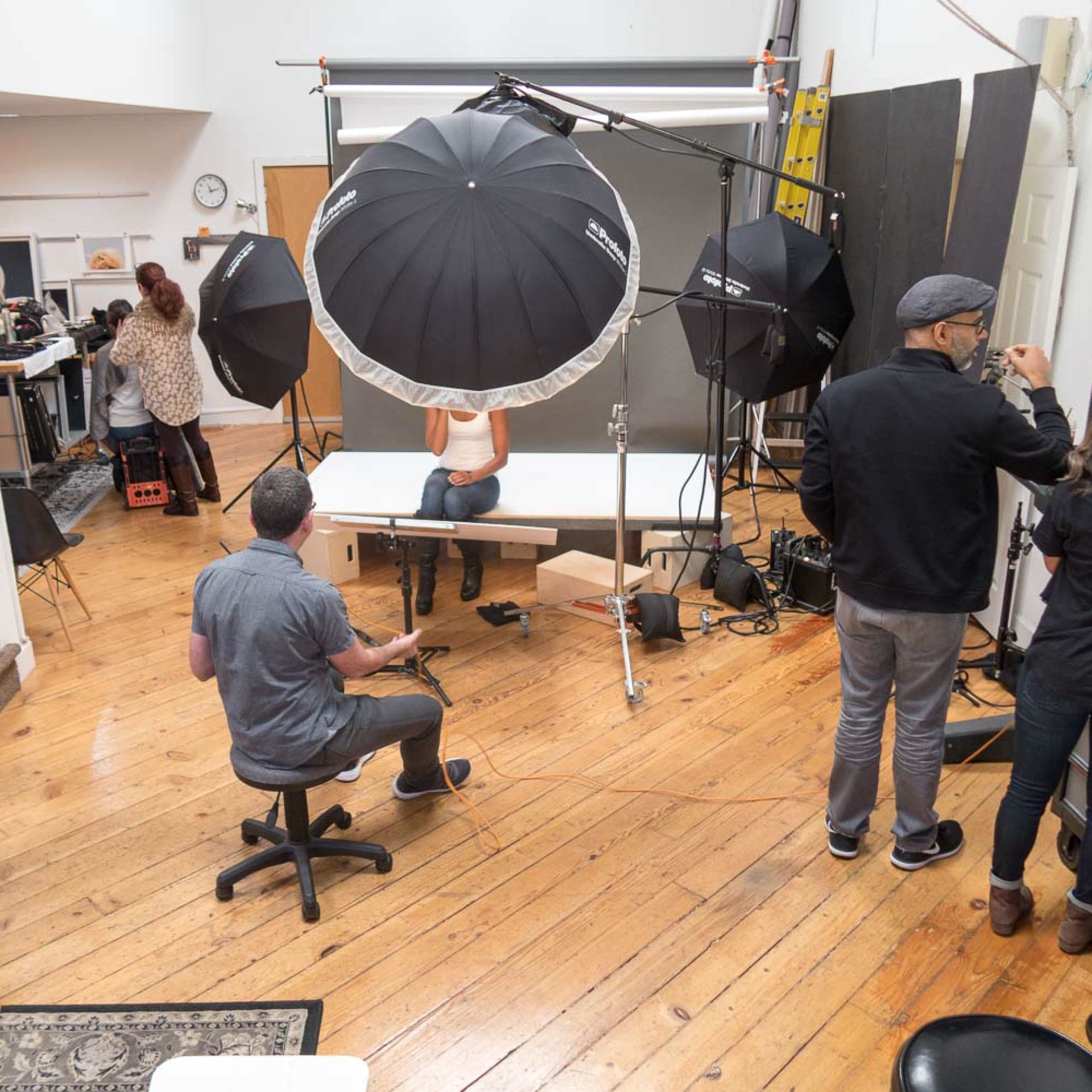 A photography studio is set up with multiple lighting equipment and a model posing on a table while several crew members assist nearby.