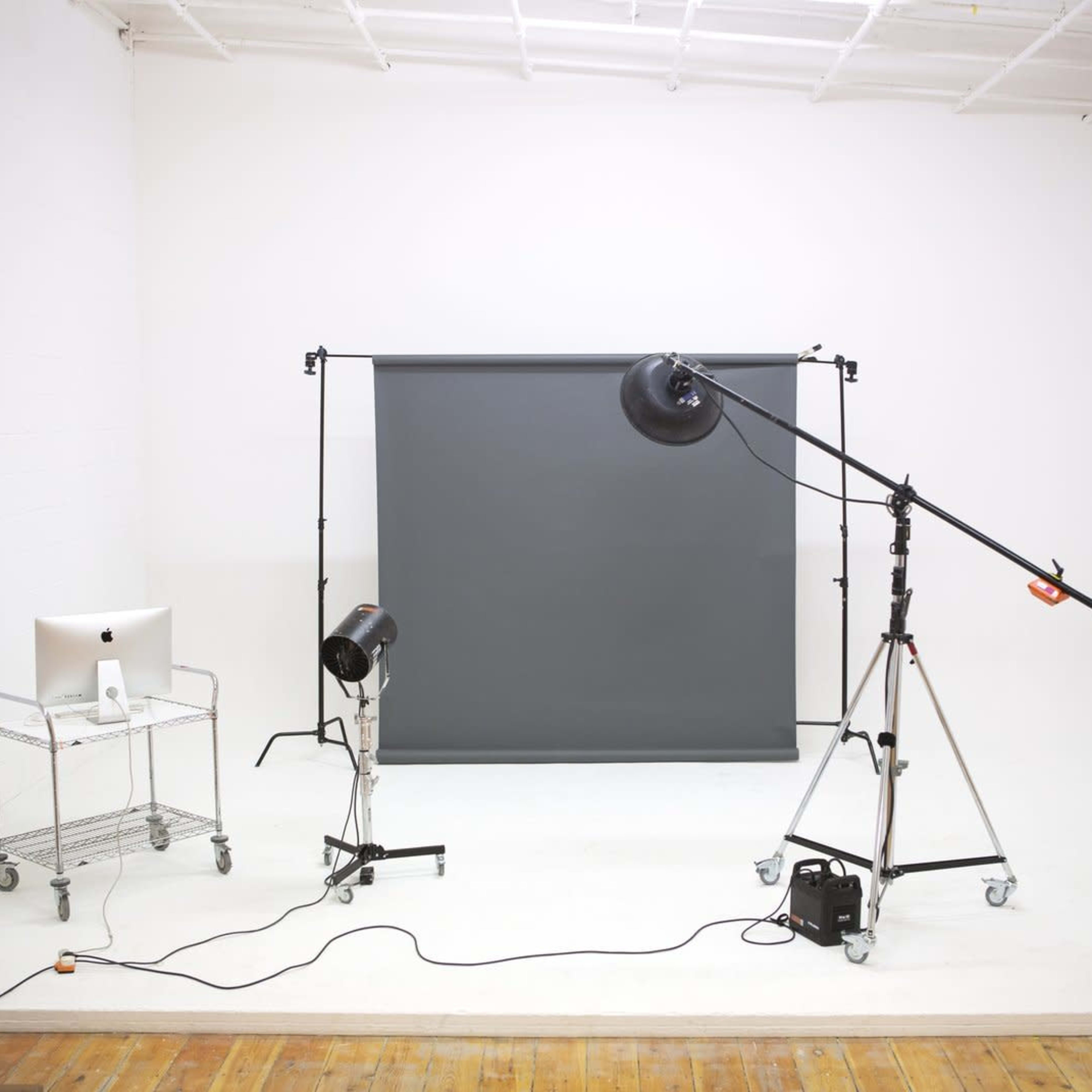 A photography studio is set up with a gray backdrop, lighting equipment on stands, and a computer on a cart.