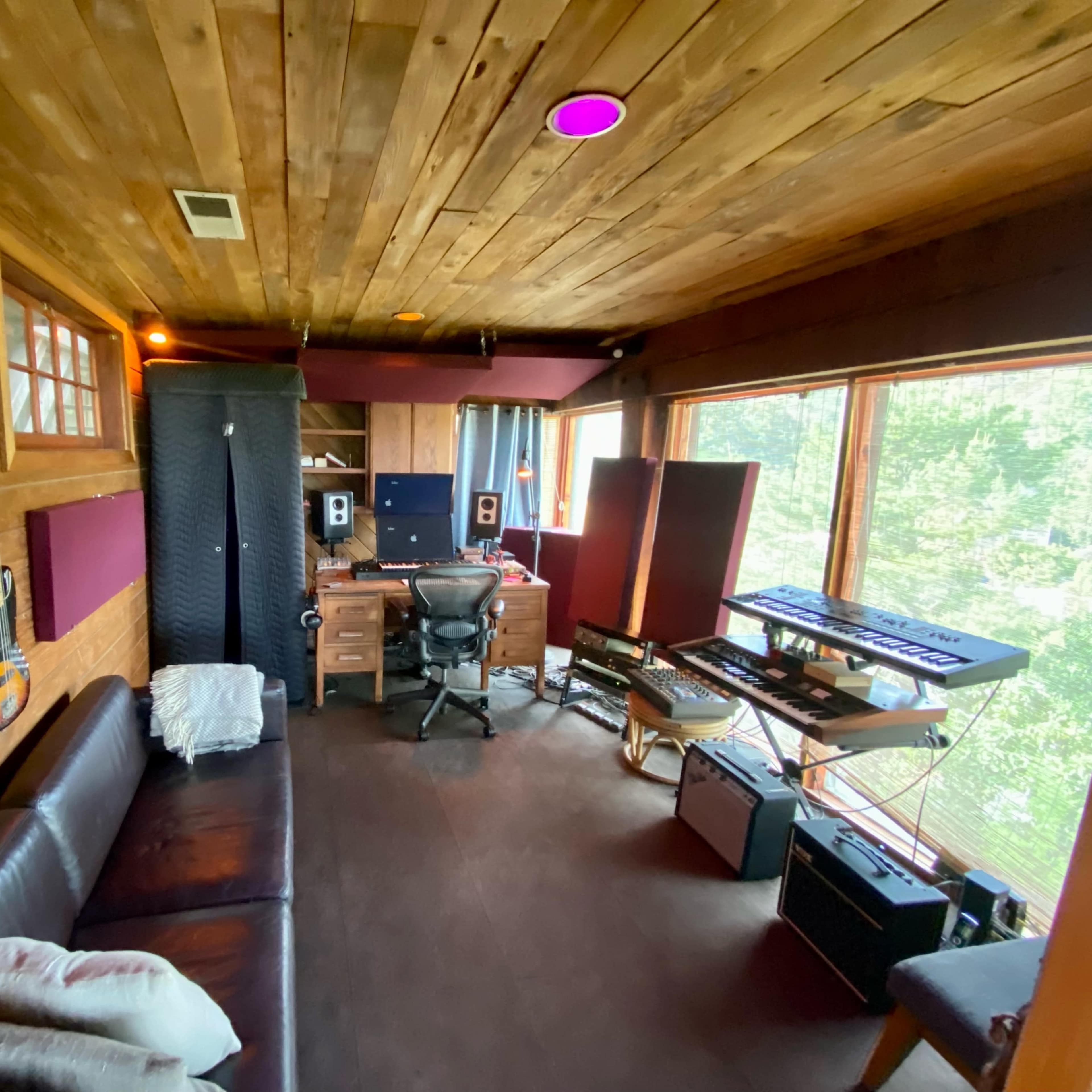 A music recording studio with wooden walls, a desk setup with audio equipment, guitars hanging on the wall, and large windows offering a view of nature.