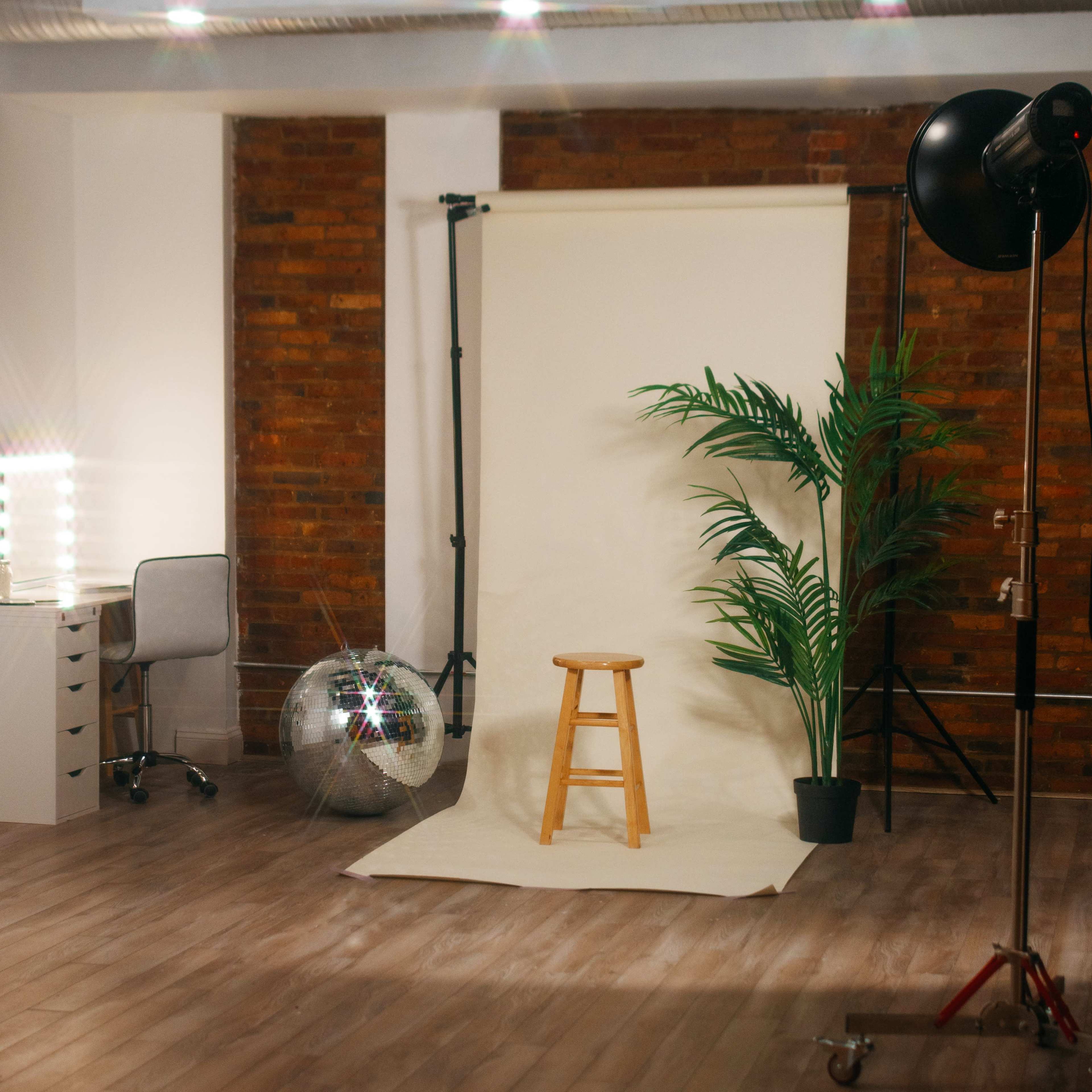 A photography studio with a wooden stool in front of a backdrop, a plant in a pot, a disco ball, and a makeup station with lights.