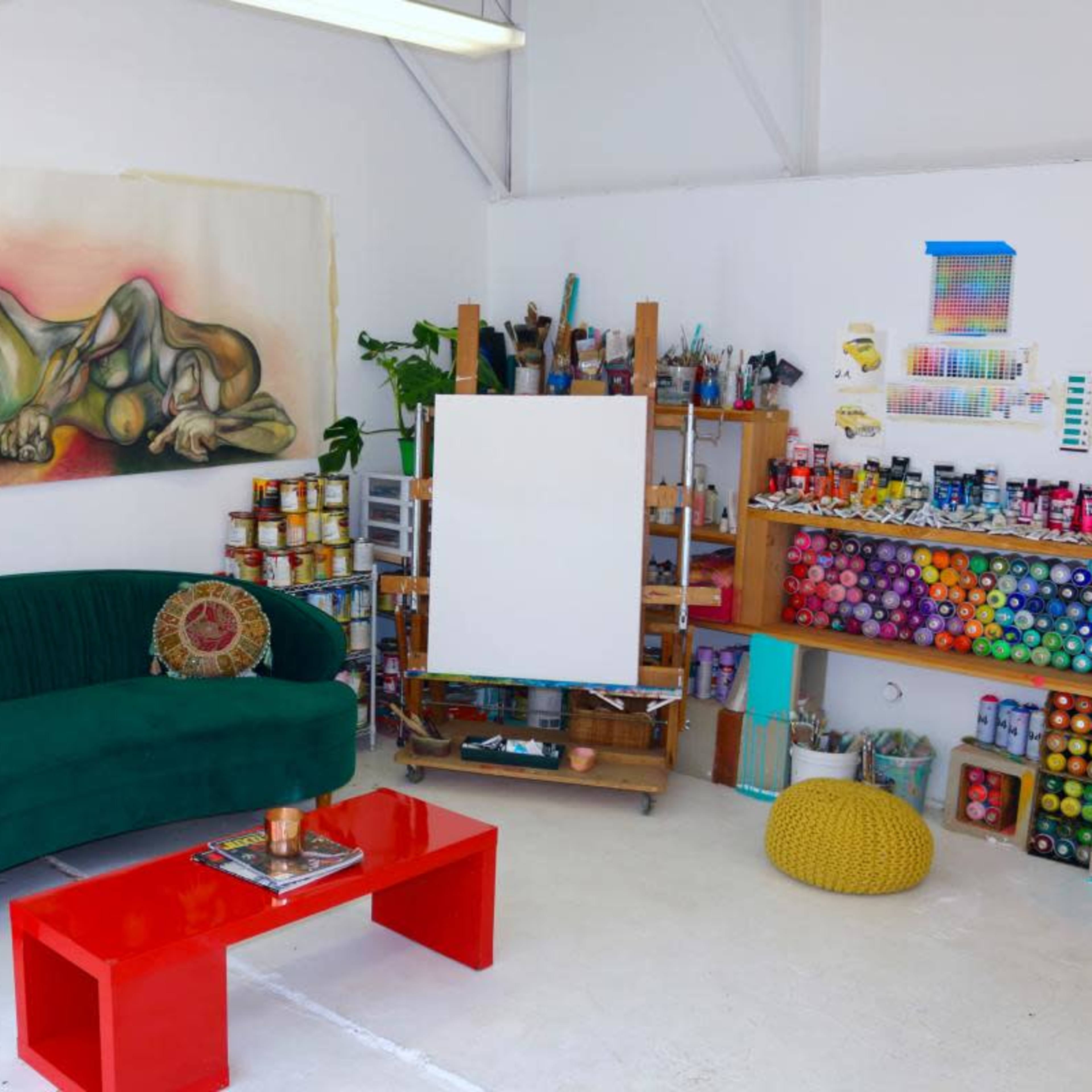 A bright art studio featuring a green velvet couch, a red coffee table, an empty canvas, and shelves stocked with various paints and art supplies.