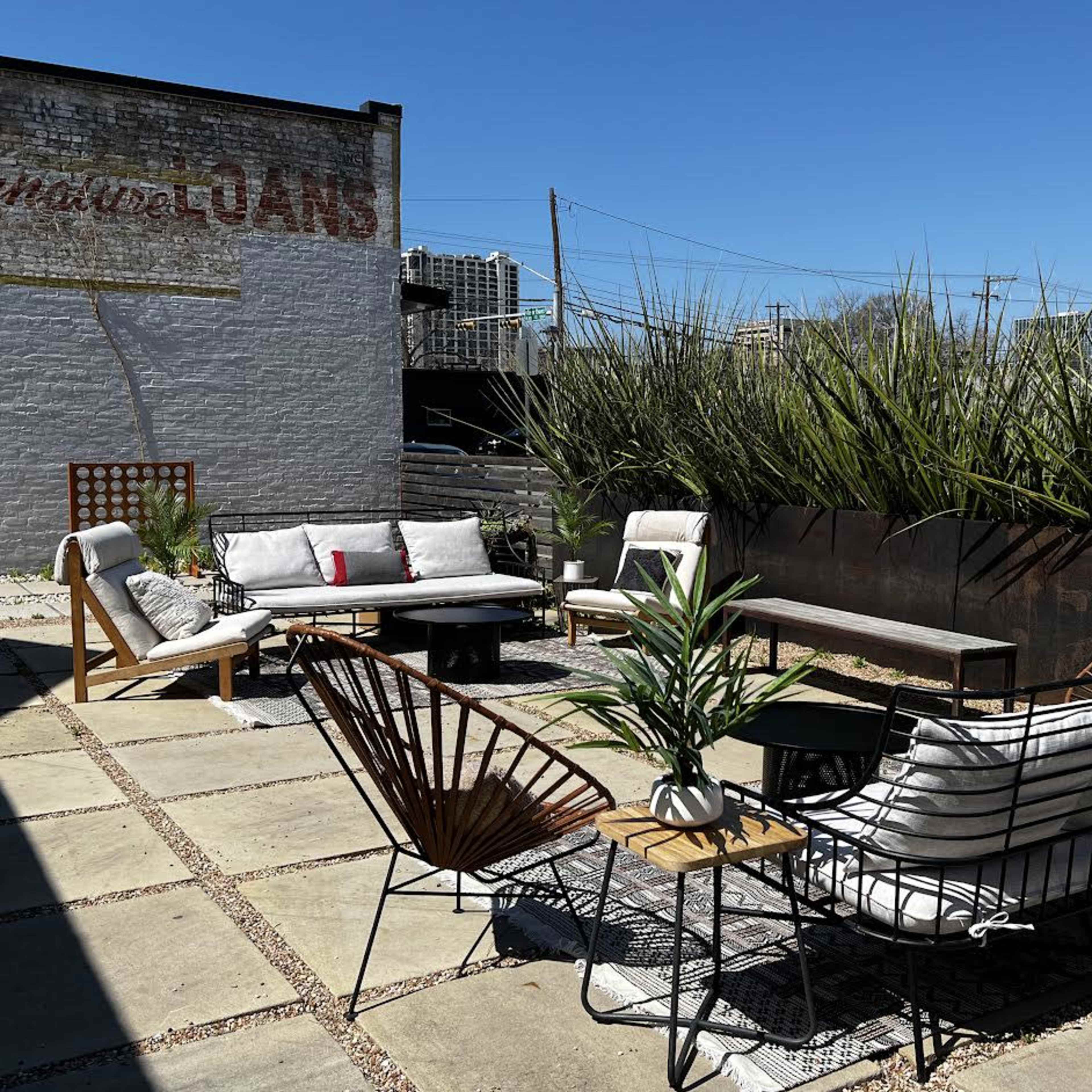 A rooftop patio with various outdoor furniture, including a sofa set and a coffee table, surrounded by tall greenery under a clear blue sky.