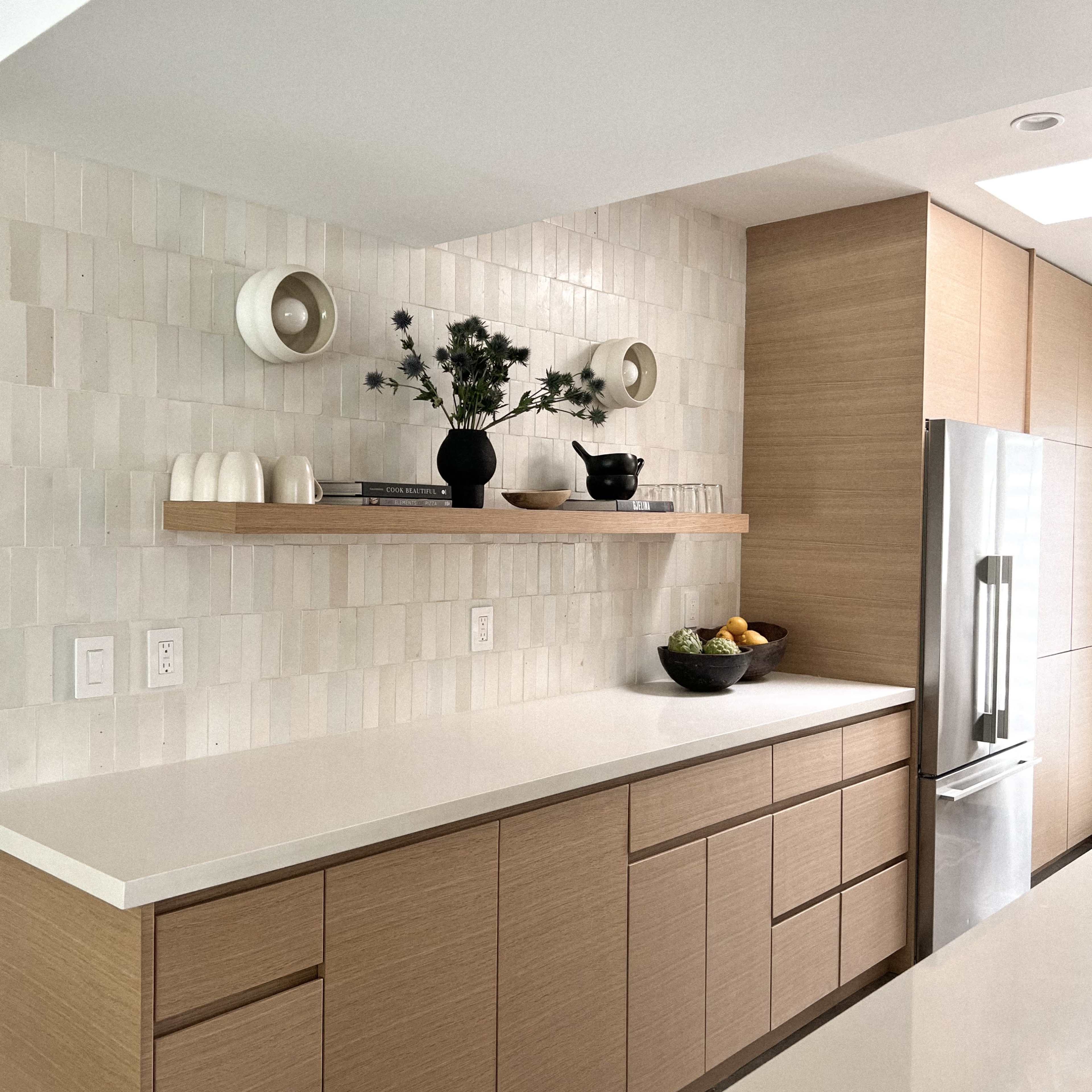 The kitchen features a light-colored countertop with a wooden base, a shelf displaying decorative items, and a bowl of fruit on the counter.