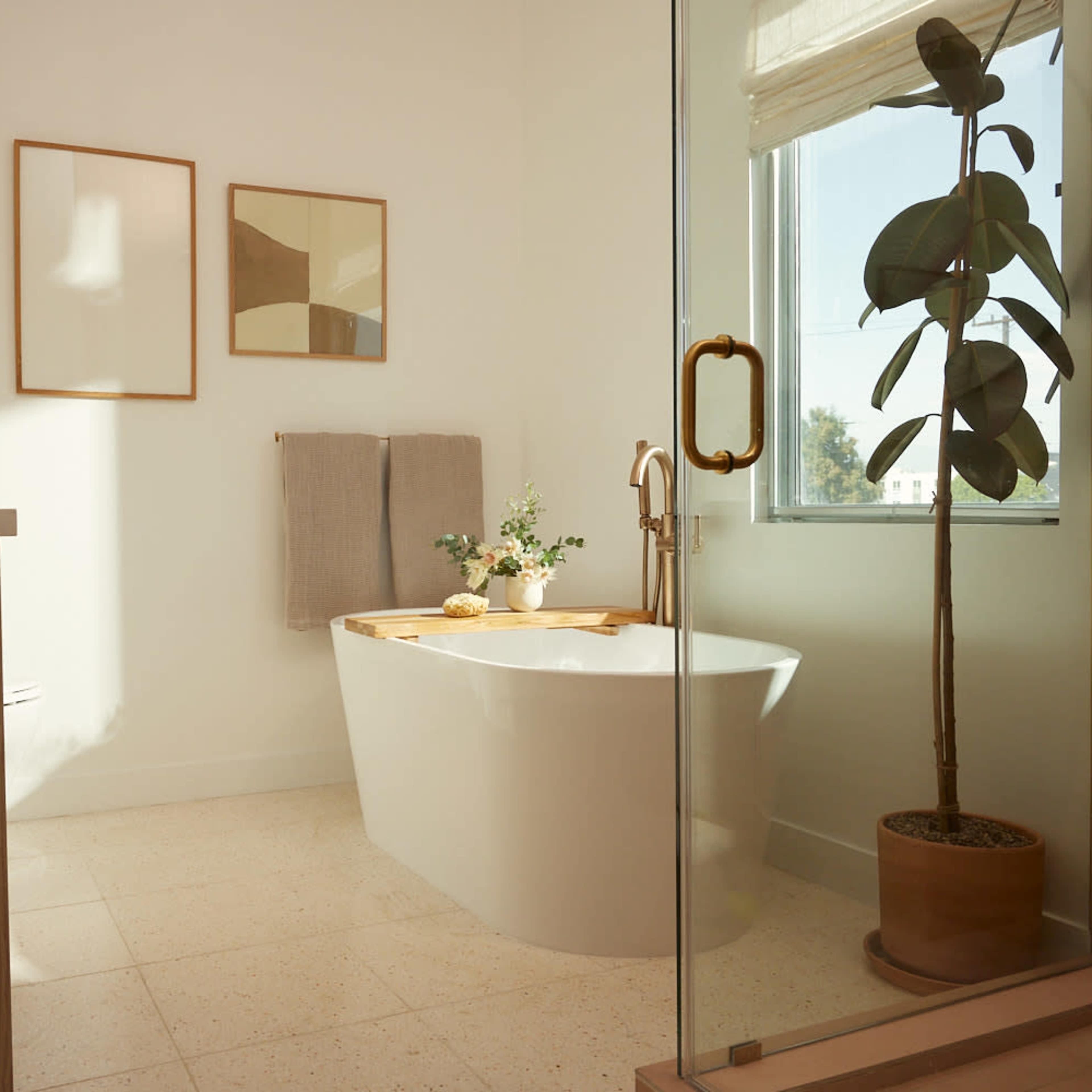 A bright bathroom featuring a freestanding tub, a wooden counter with a stool, two framed artworks, and a potted plant near a window.