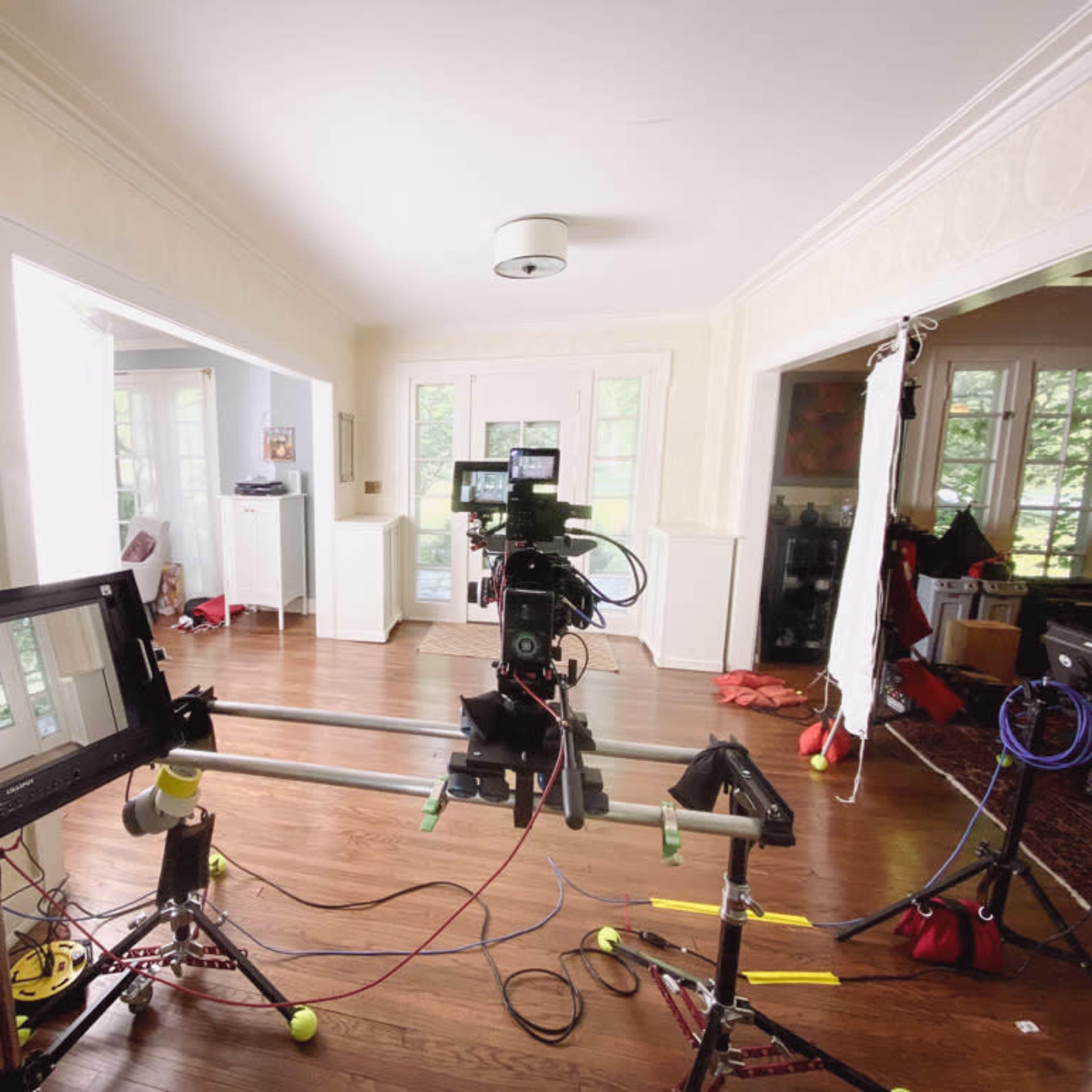 A film setup in a spacious room featuring a camera on a slider track, with monitors and lighting equipment arranged around a bright, open living area.