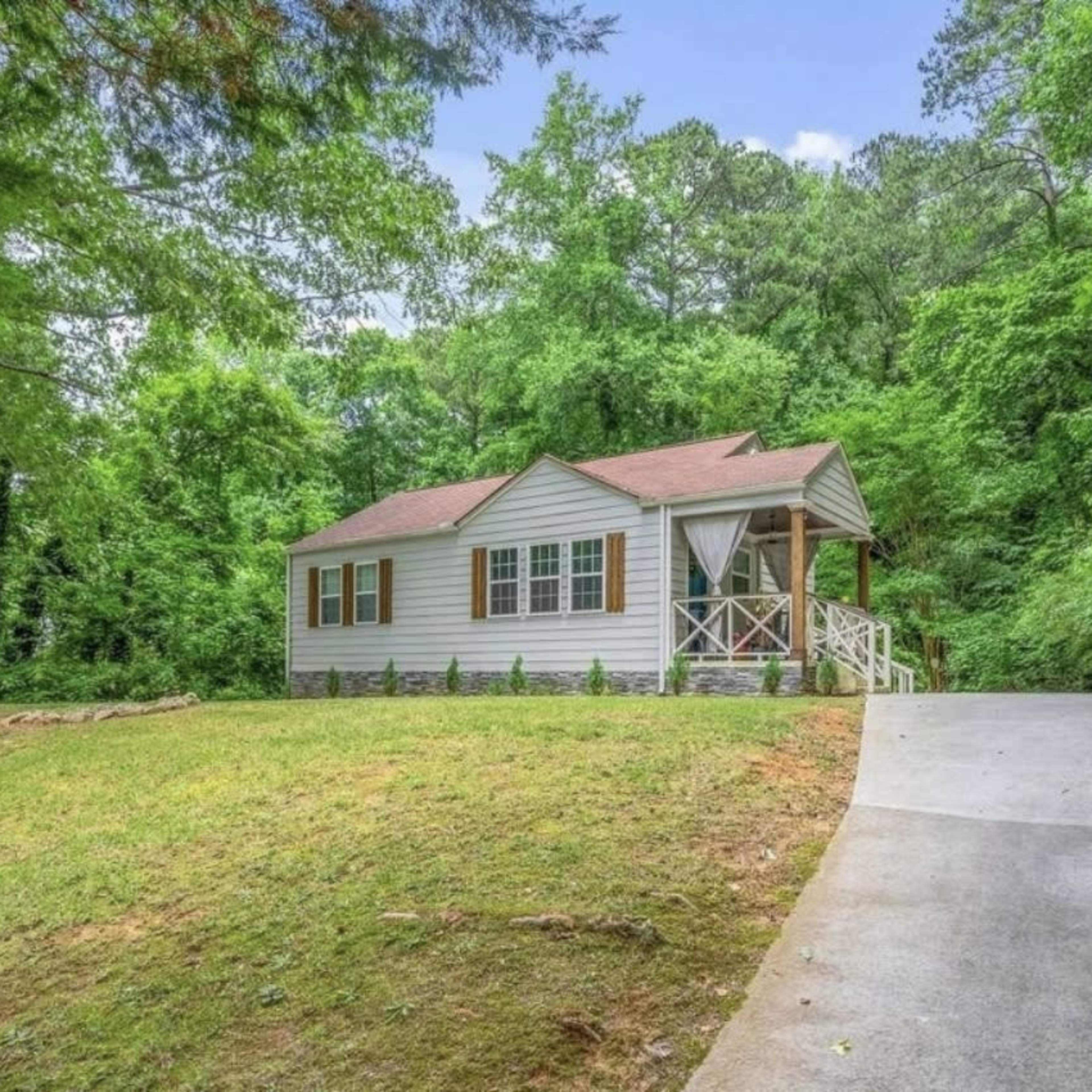 A small, single-story house with a sloped roof and front steps is situated on a grassy area surrounded by trees.