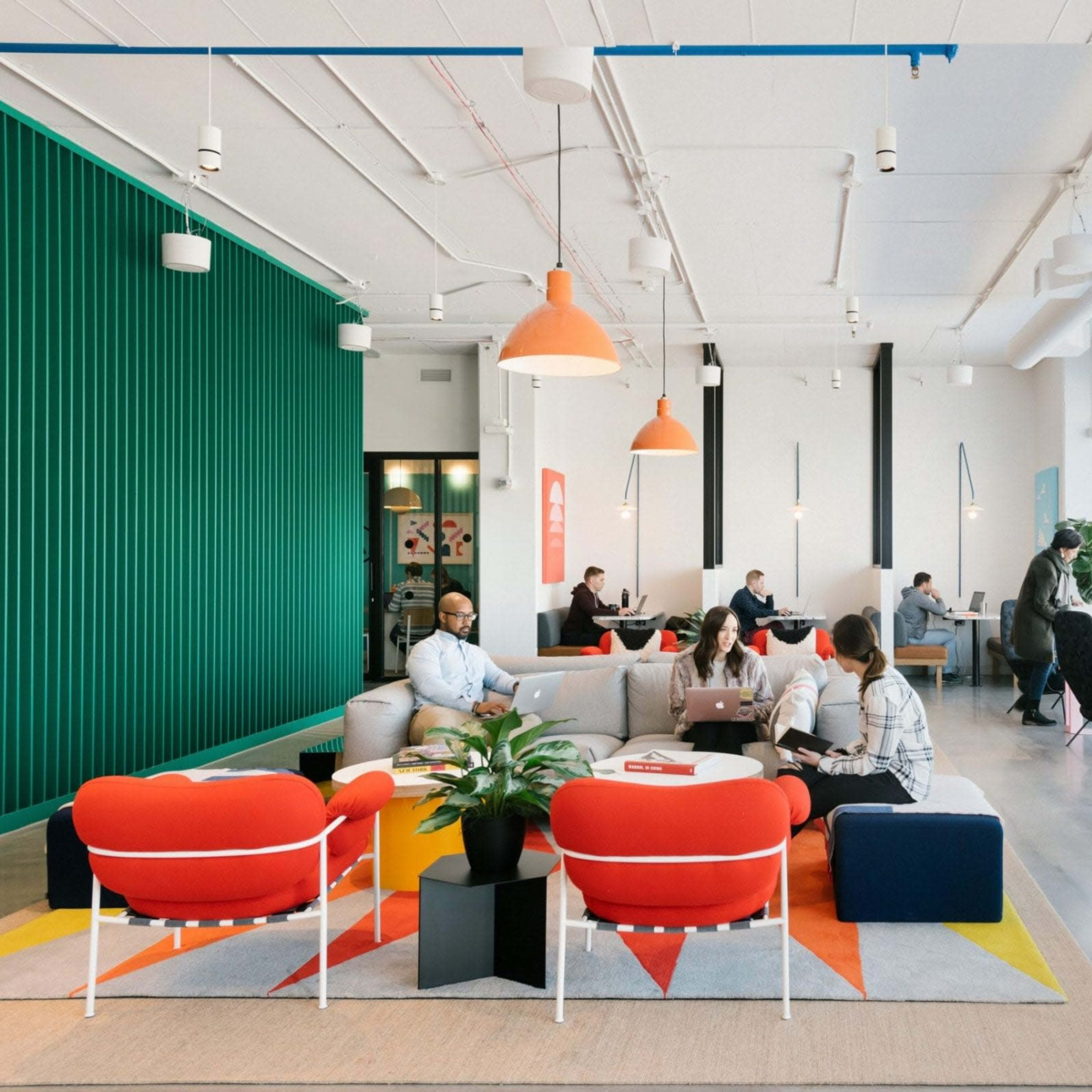A modern office lounge area with colorful seating, large windows, and several people working and socializing.