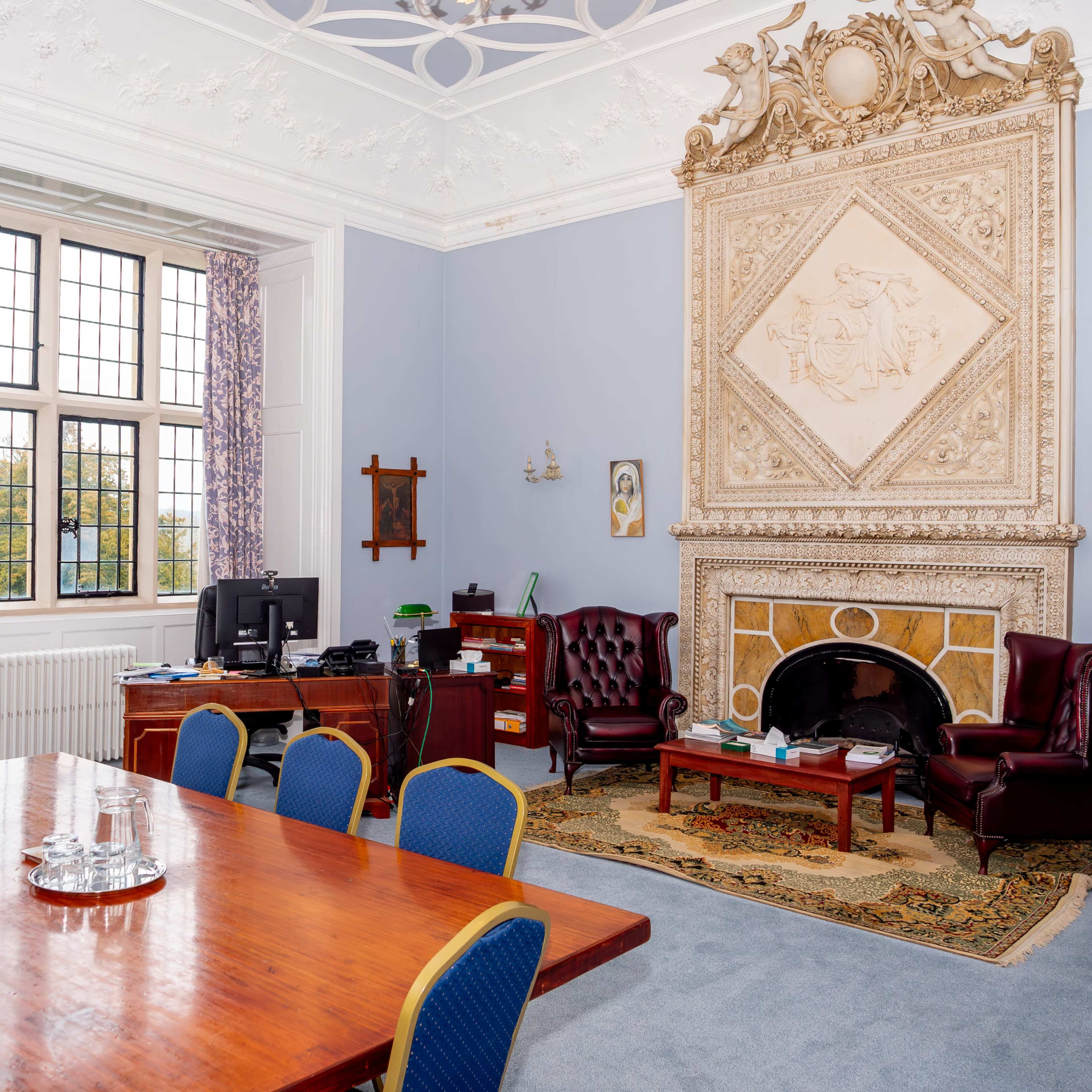 A spacious room with a large wooden table, two armchairs by a fireplace, and a desk equipped with a computer.