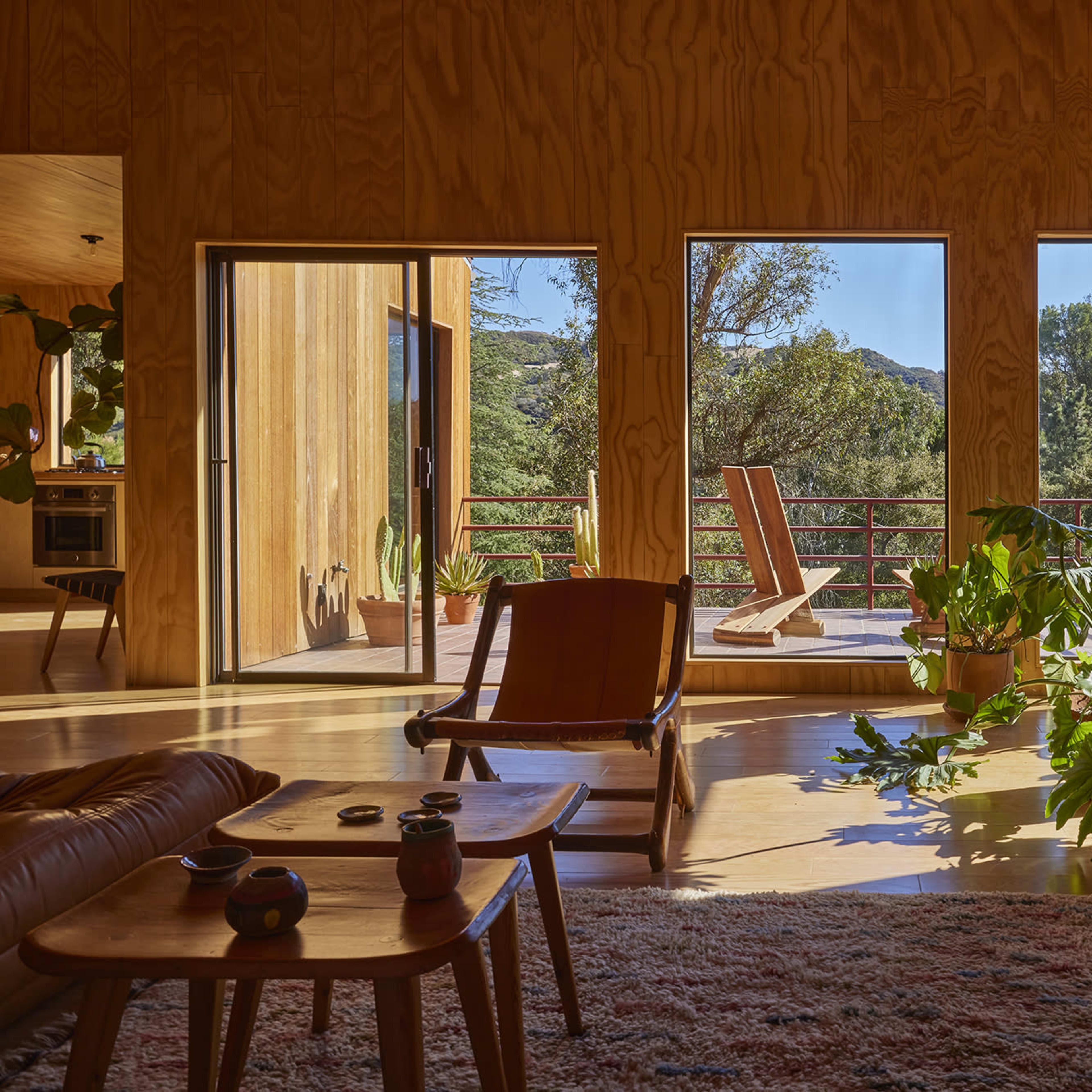 The interior of a wooden house features large windows with a view of greenery, a lounge chair, and potted plants.