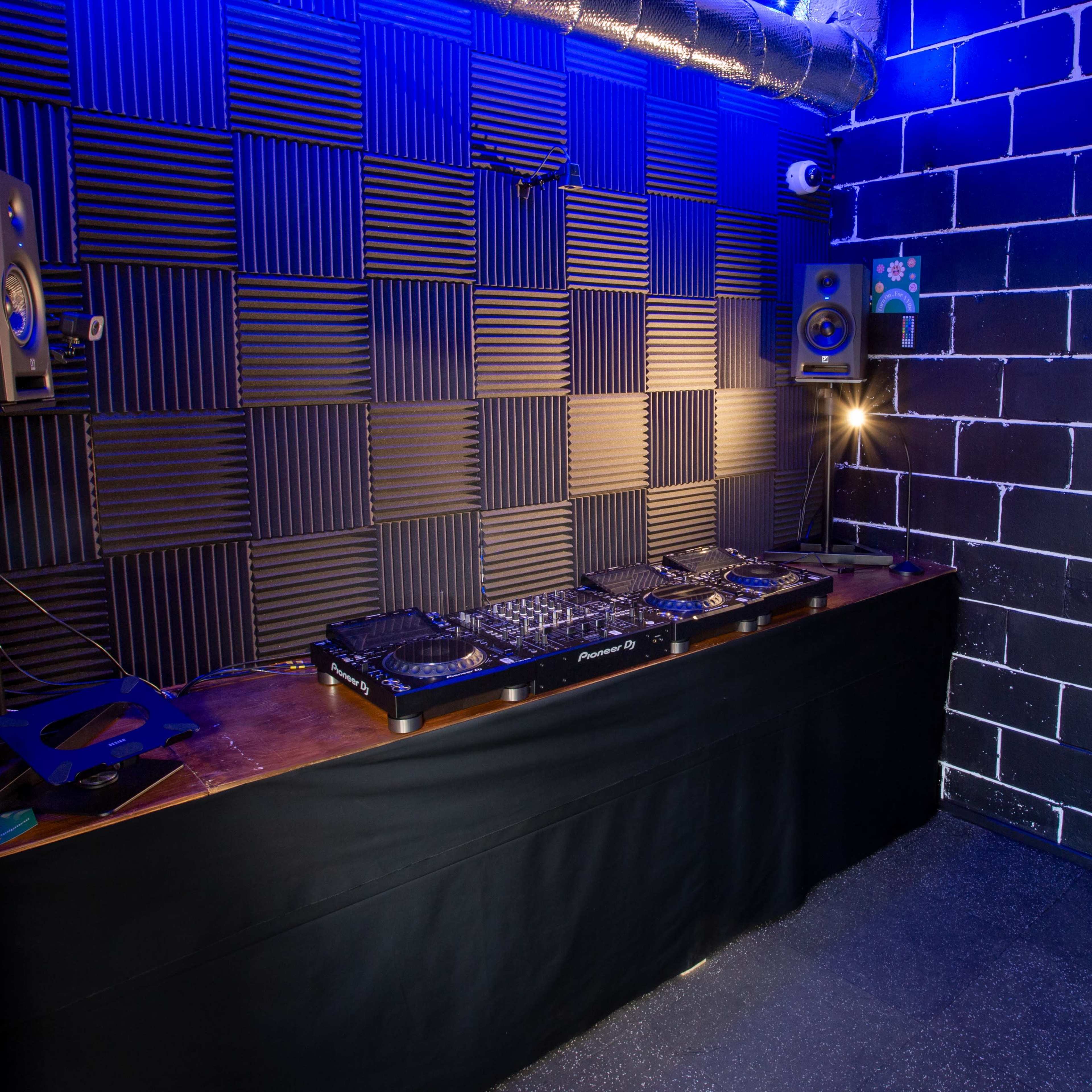 A DJ booth with a mixer and speakers in a soundproof room featuring black padded walls and dim lighting.