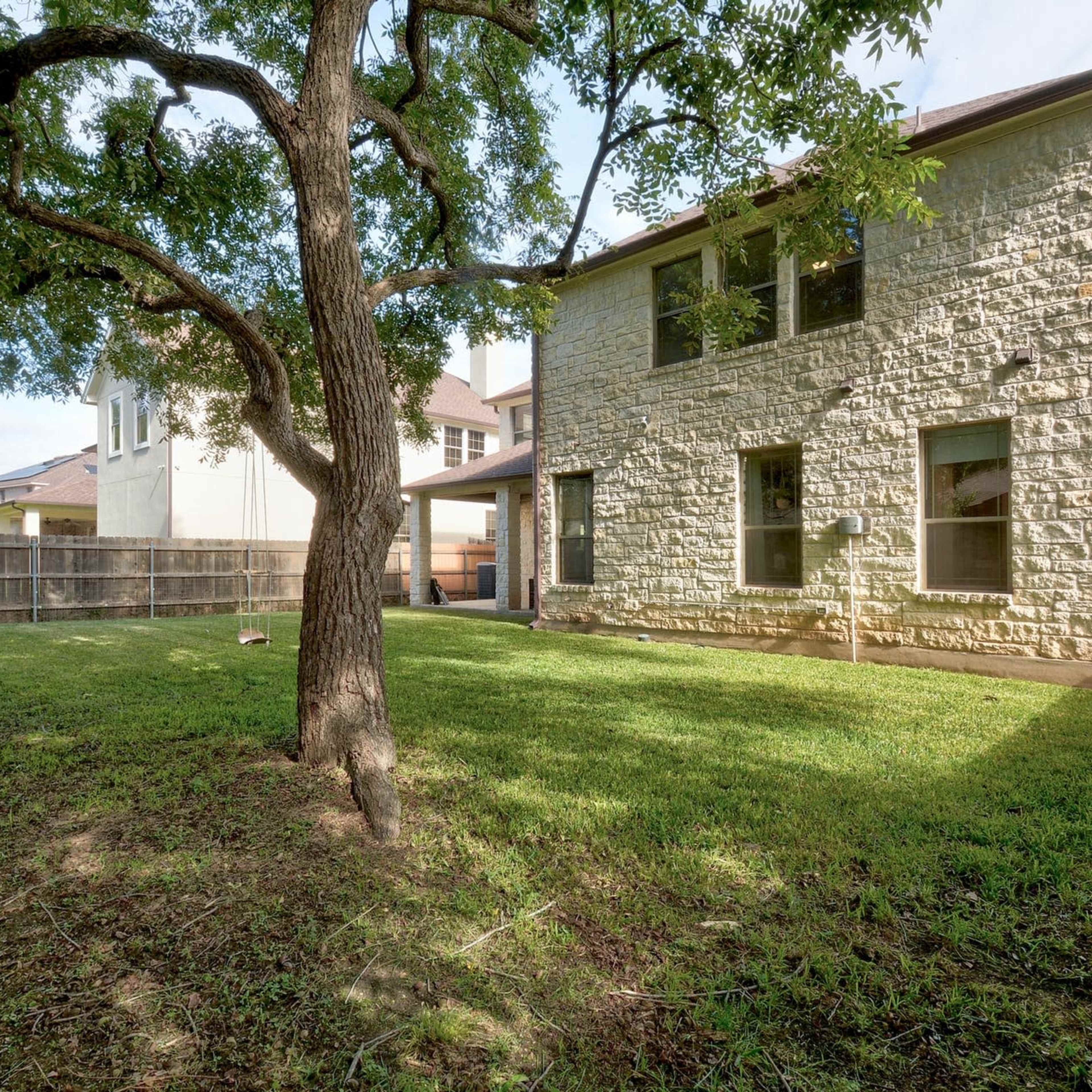 The rear view of a two-story stone house with a large grassy yard and a tree in the foreground.