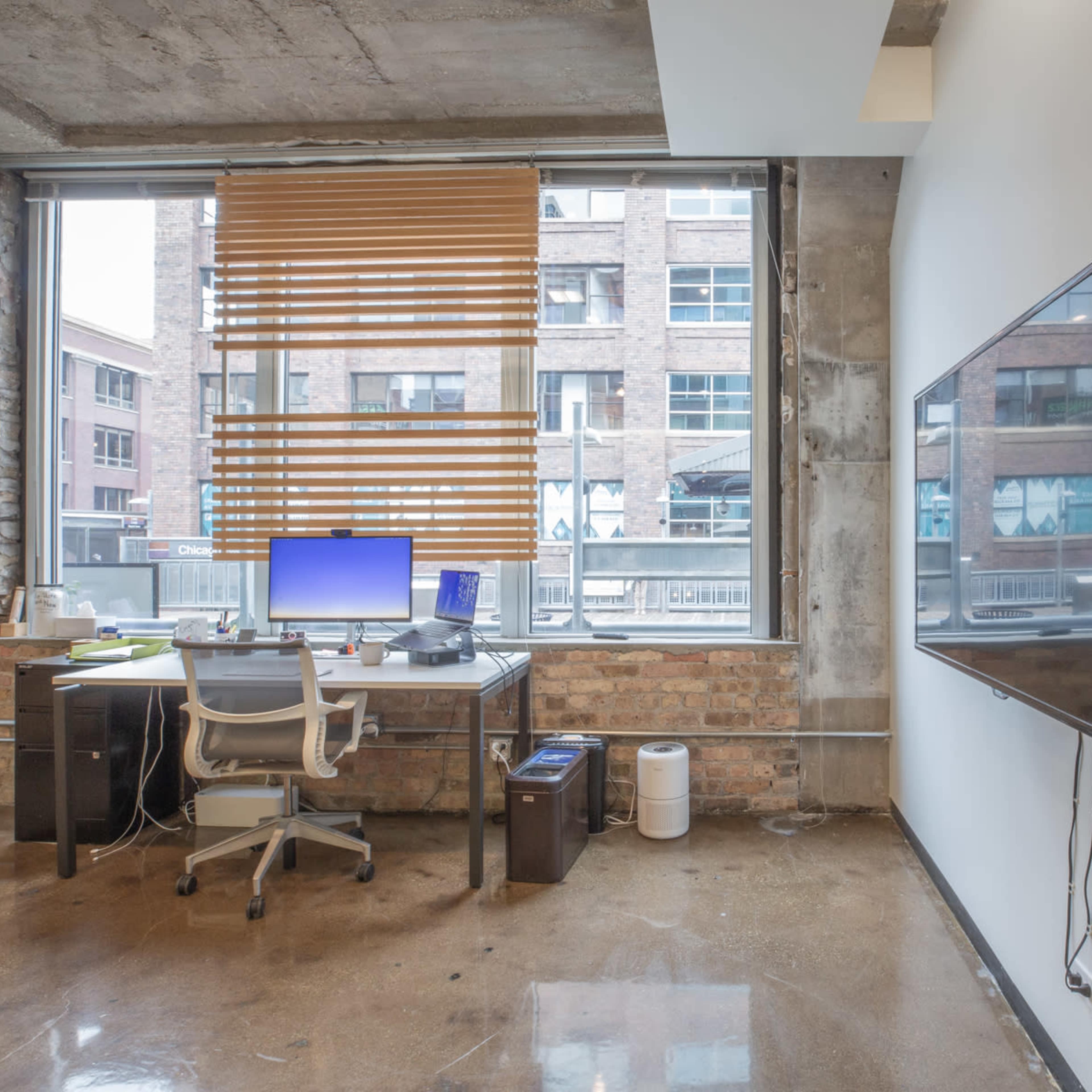 A modern office space with a desk, computer, and large window featuring wooden blinds, along with a wall-mounted television.