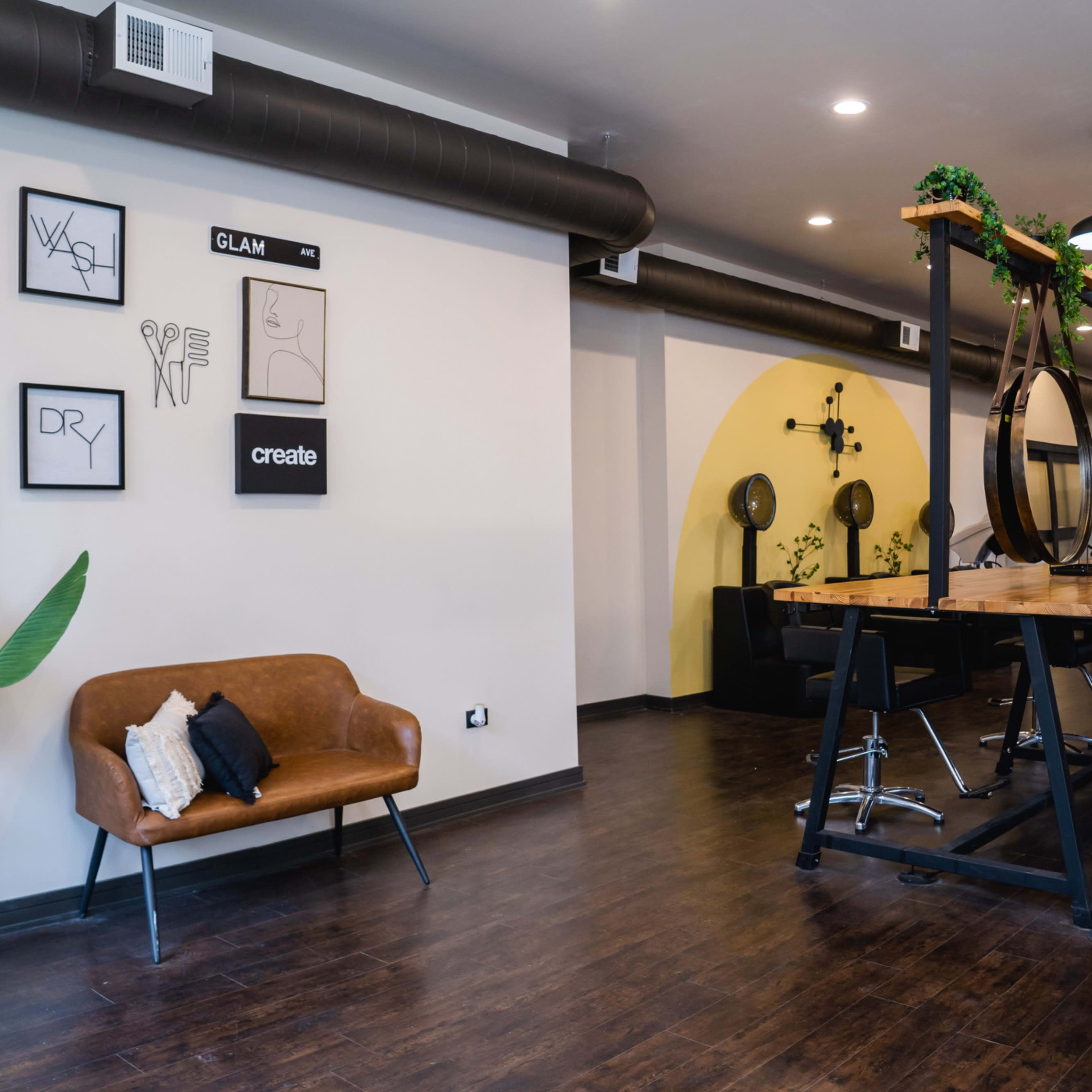 A modern salon interior with a long wooden table, black chairs, large round mirrors, and greenery on the walls.