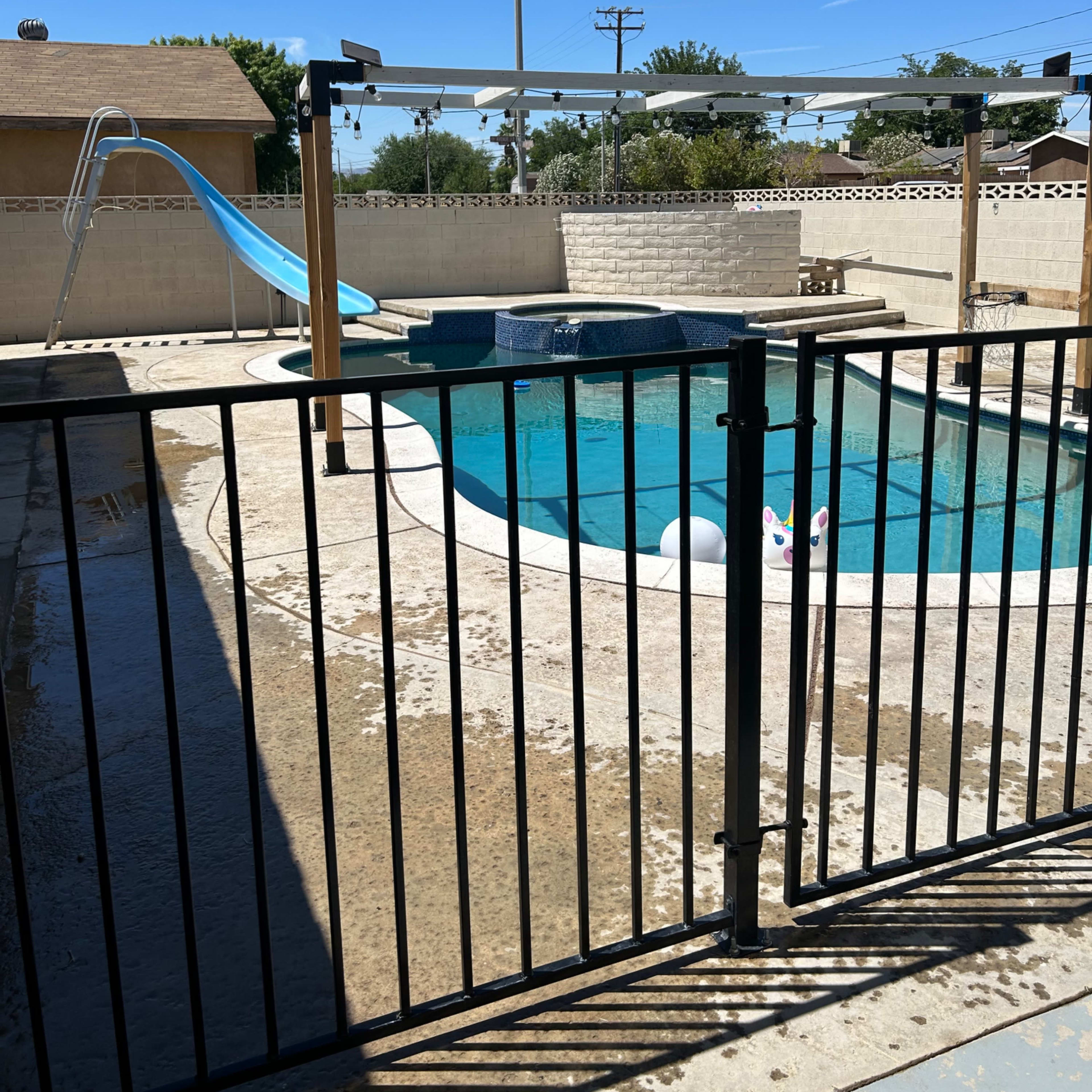 A swimming pool enclosed by a black fence, featuring a slide and a white float, under a clear blue sky.