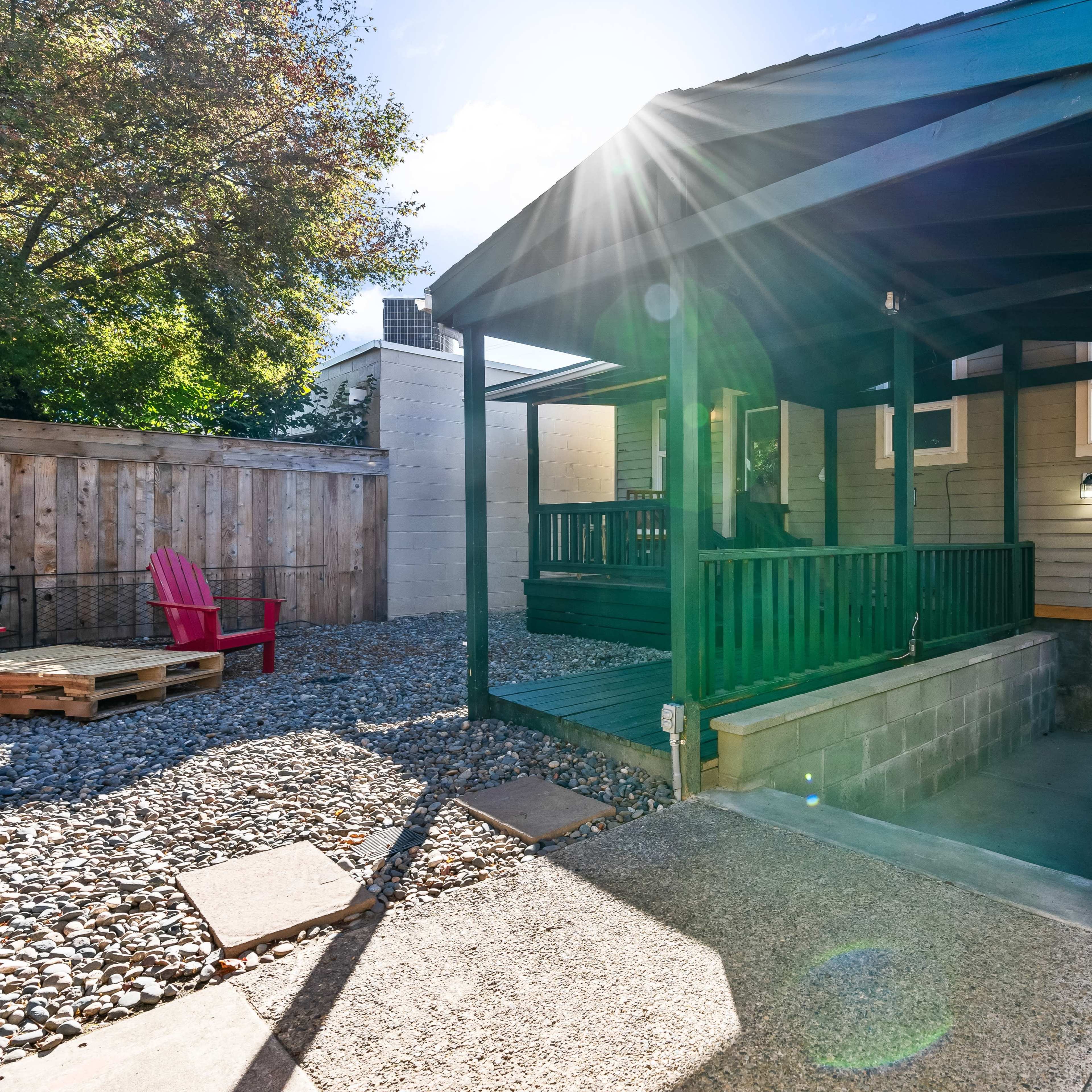 A green porch with a slanted roof is attached to a house, surrounded by a gravel yard and a red chair nearby.