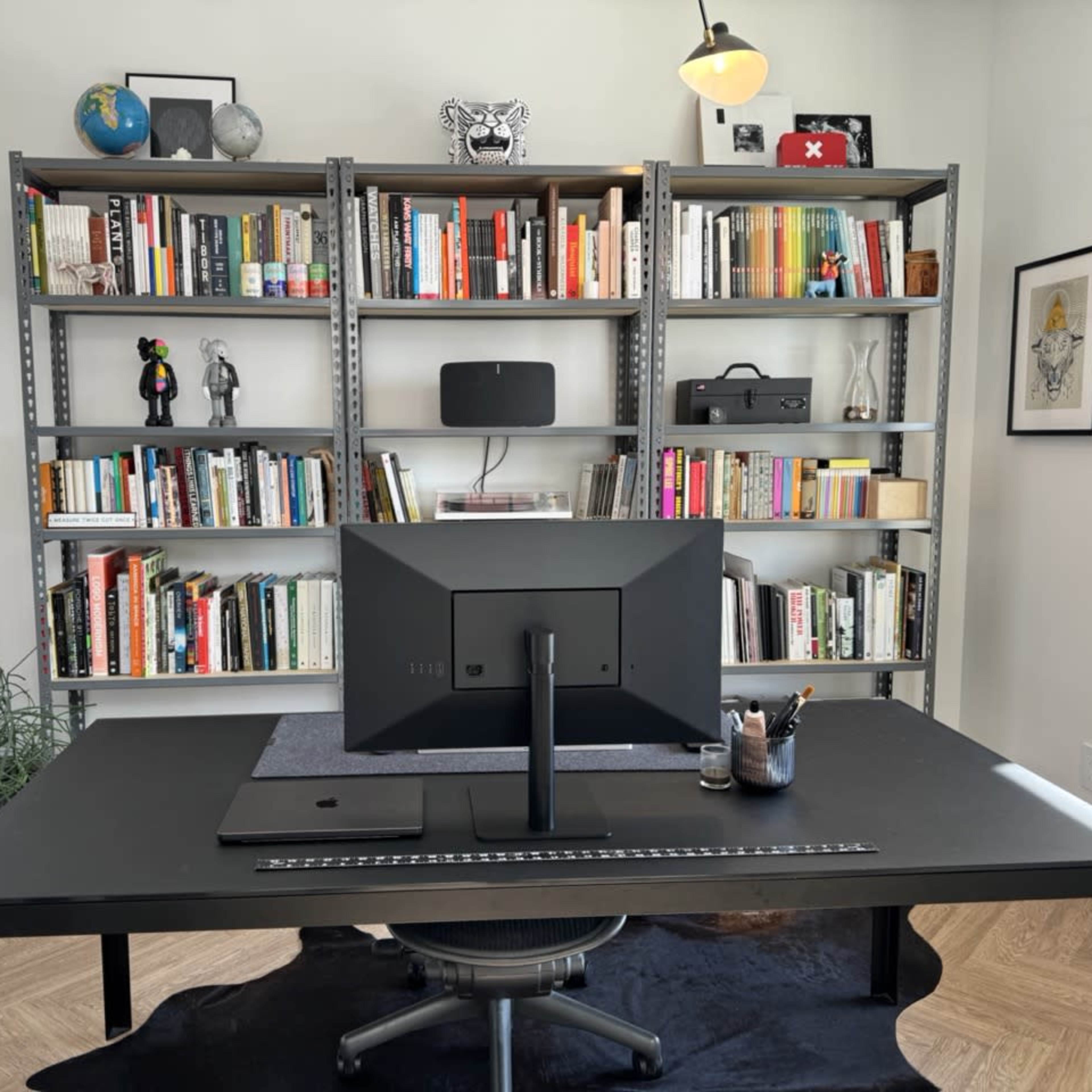 A modern workspace features a black desk with a computer monitor, surrounded by bookshelves filled with books and decorative items.